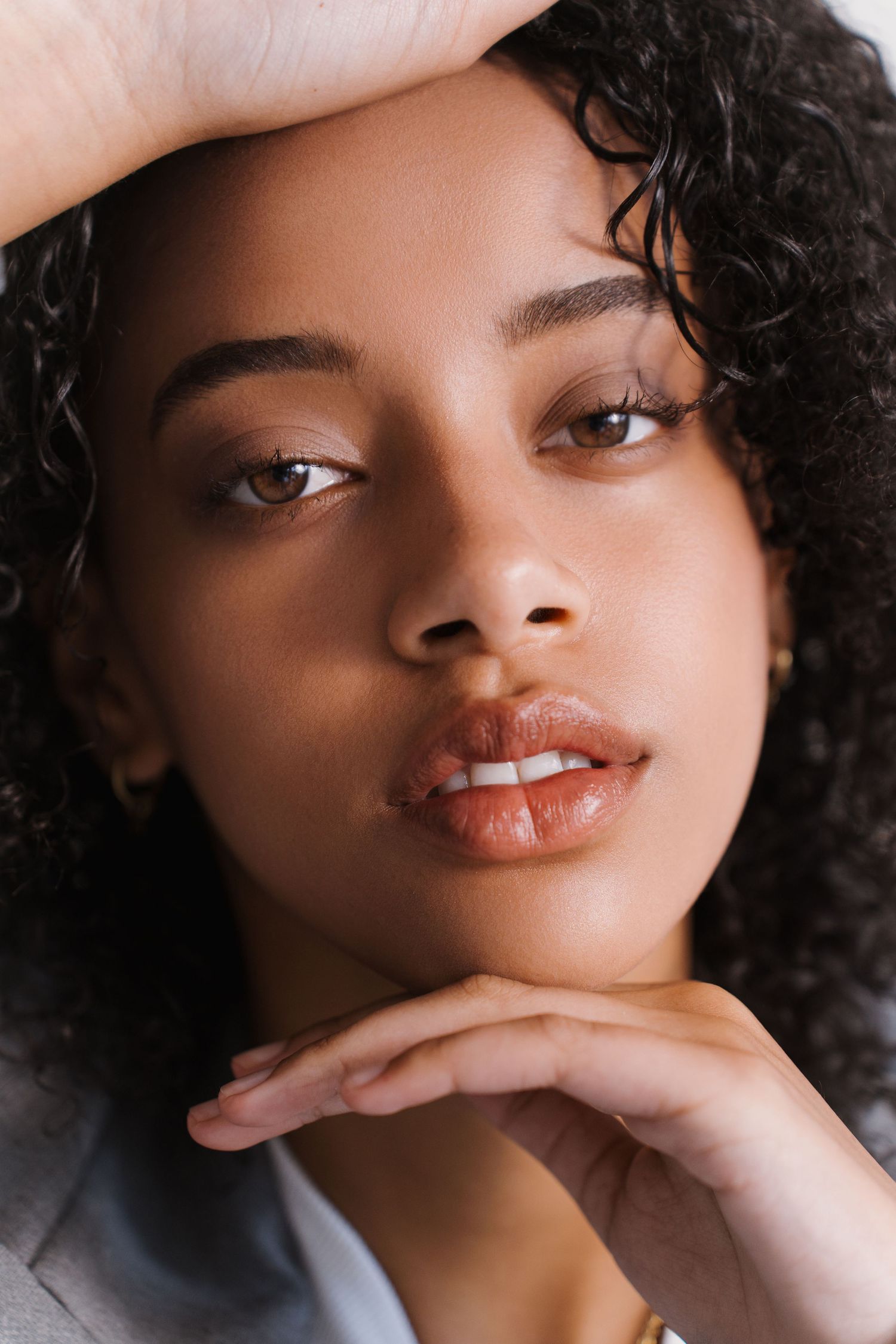 Close-up portrait of a young woman with curly hair resting her chin on her hand against a white background.