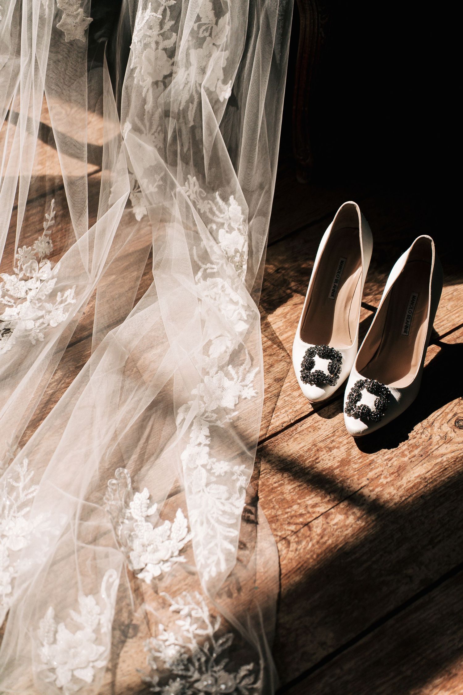 Dramatic sunlit flat lay of intricate lace wedding dress hem beside white bridal heels with black embellished buckle on wood floor.