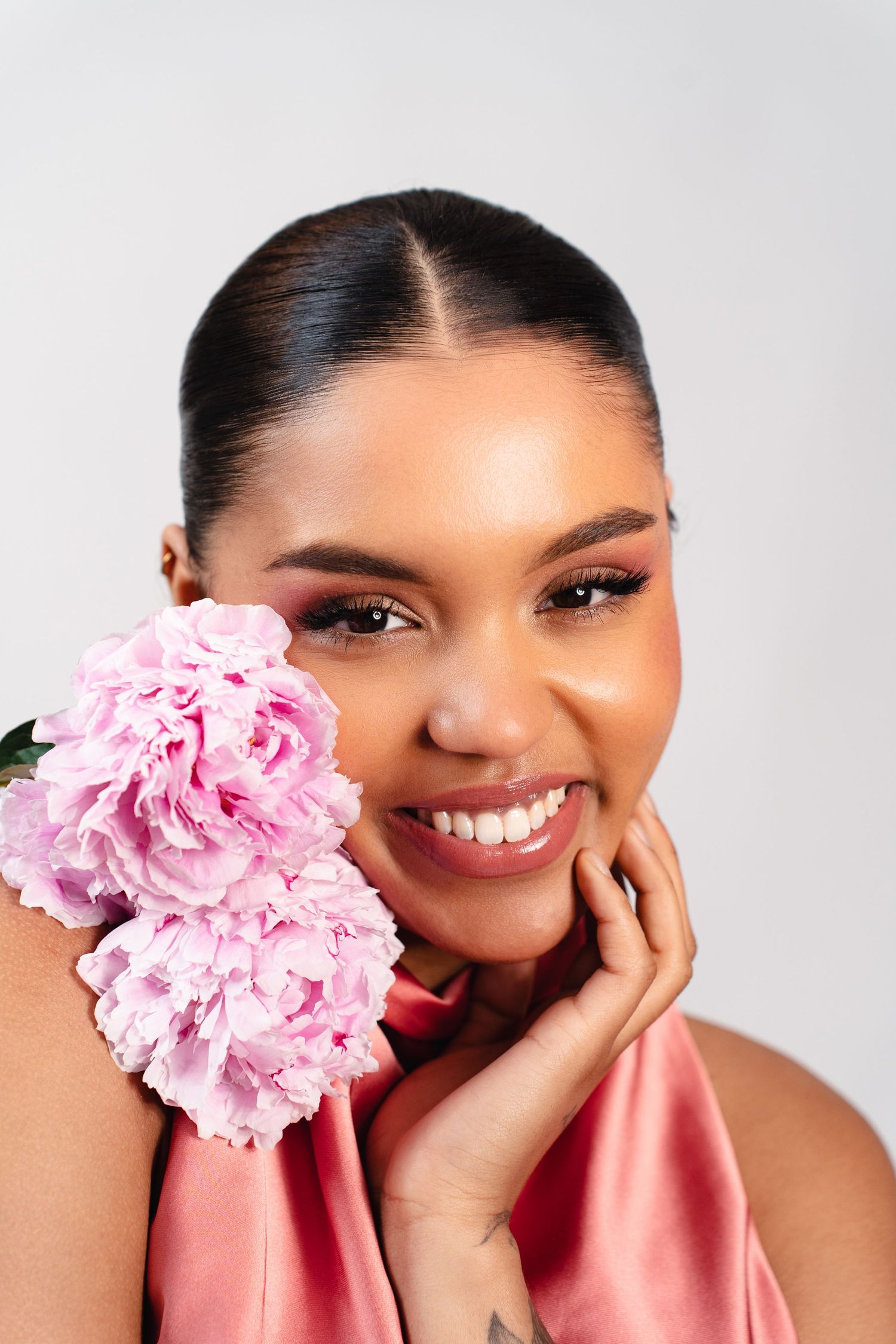 Smiling woman in pink satin top holding a pink peony flower beside her face against a light gray background.