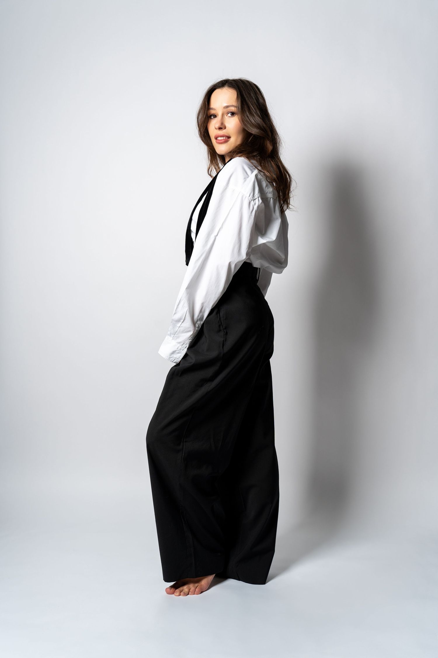 Woman in white shirt and black wide-leg pants posing sideways glancing back in studio portrait.