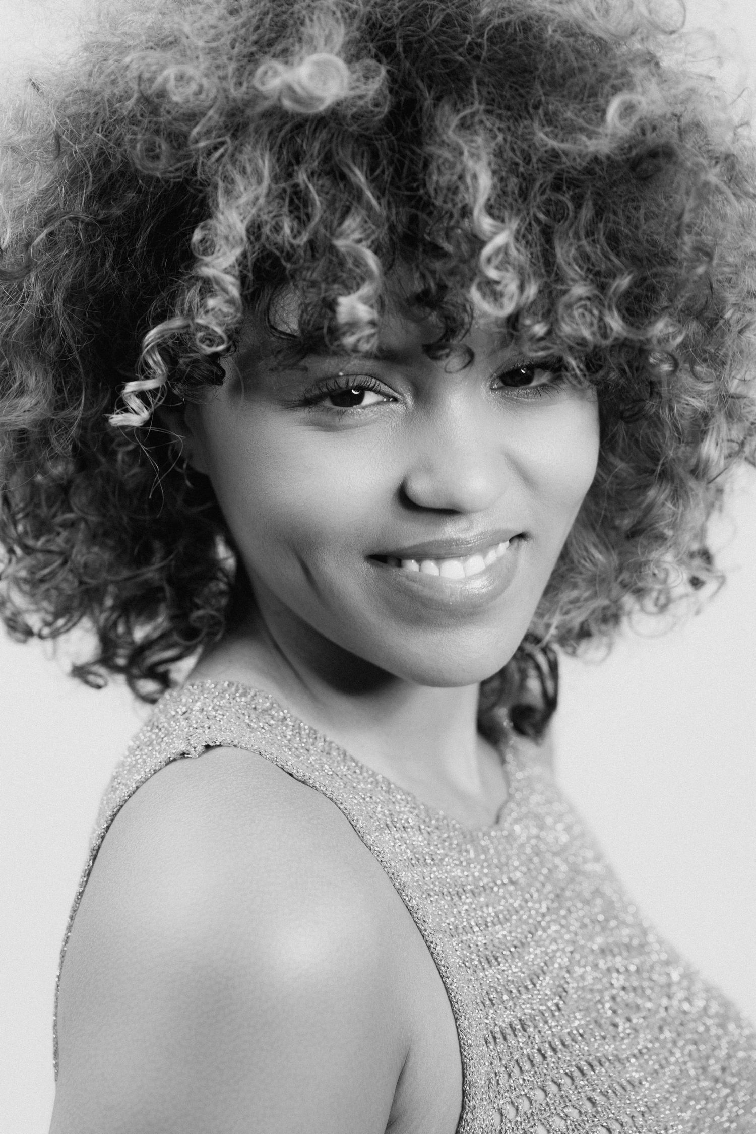 Black and white close-up portrait of a young woman with voluminous curly hair smiling over her shoulder.