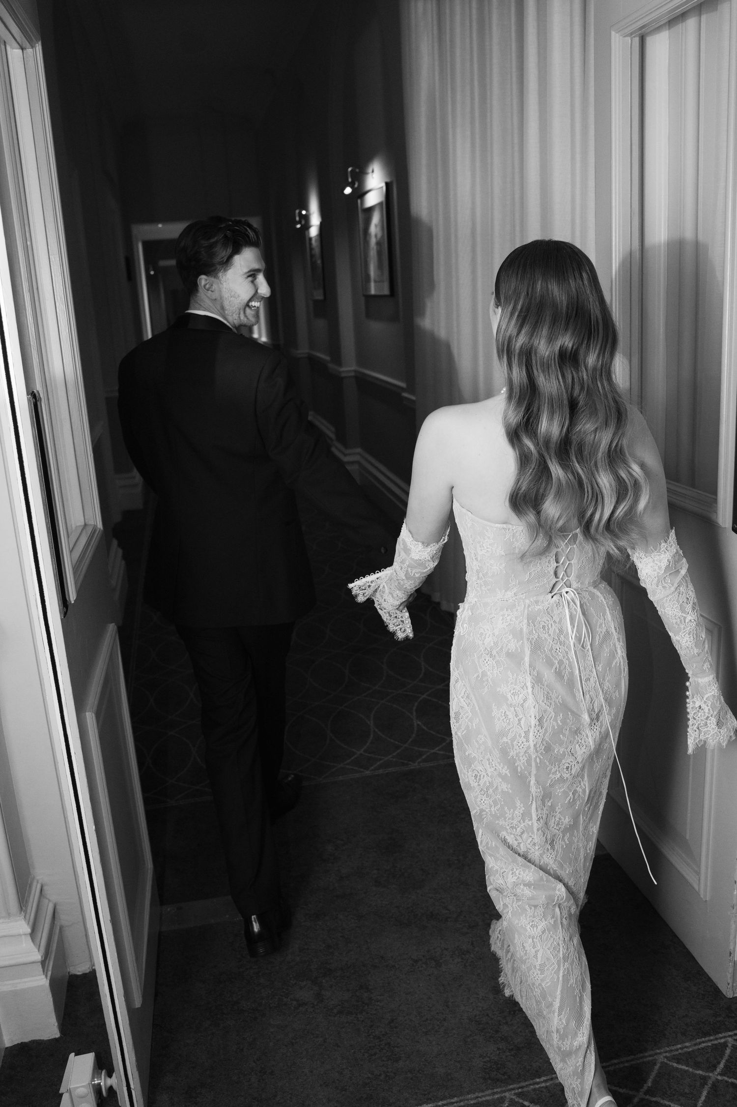 Black and white photo of bride and groom holding hands while walking through a grand hotel corridor.