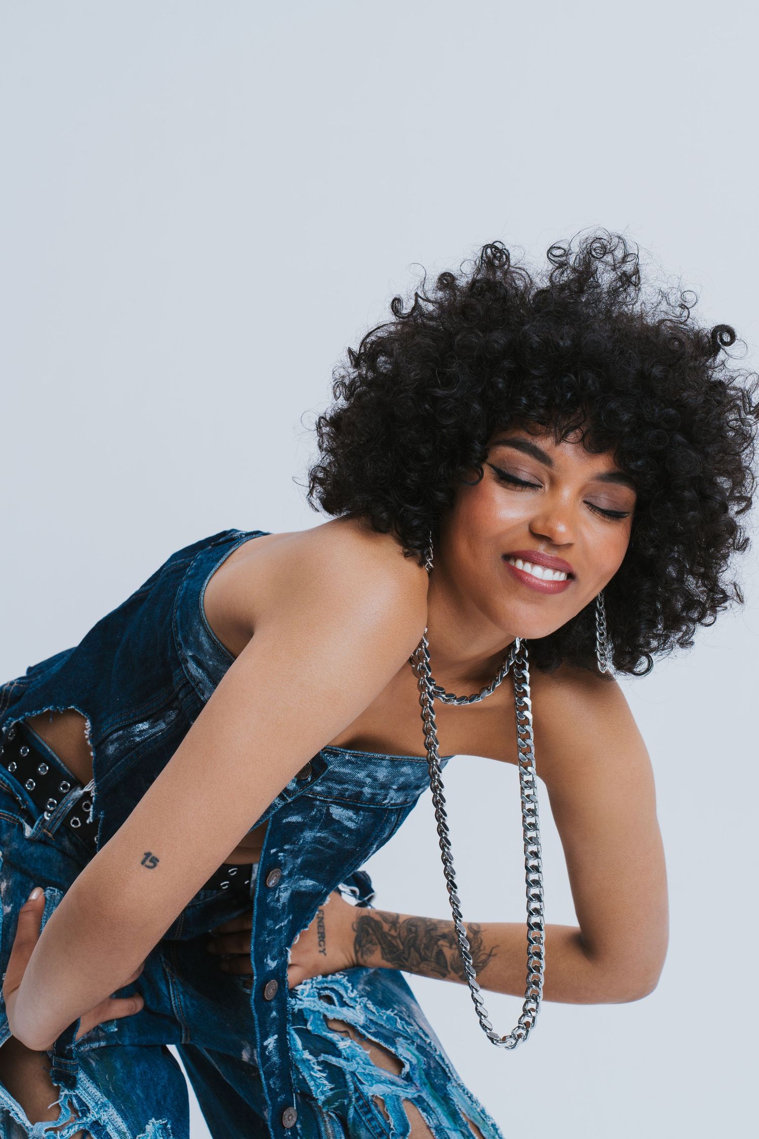 Young woman with curly afro hair smiling, wearing denim outfit and silver chain necklaces in studio.