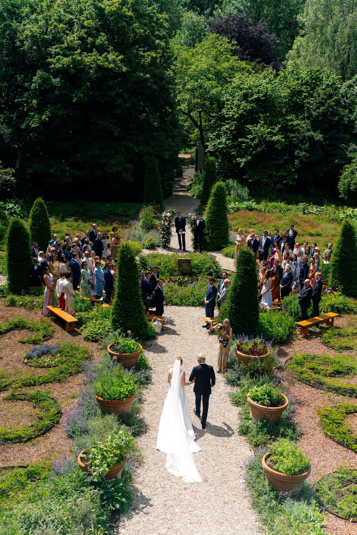 Aerial view of a bride walking down a garden aisle with lush greenery, topiary trees, and wedding guests lining the path.