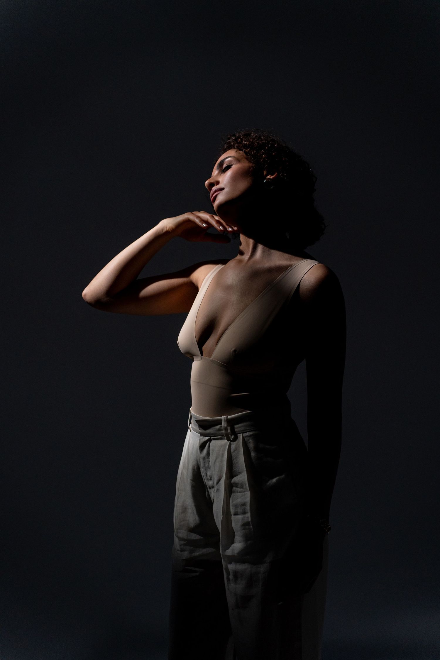 Dramatic dark portrait of a woman in a bodysuit and trousers touching her neck gracefully.