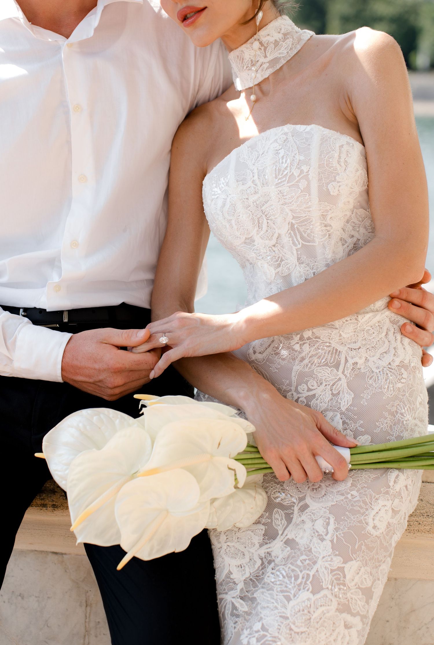 Bride in lace wedding gown holding white calla lily bouquet beside groom in white shirt and black trousers.