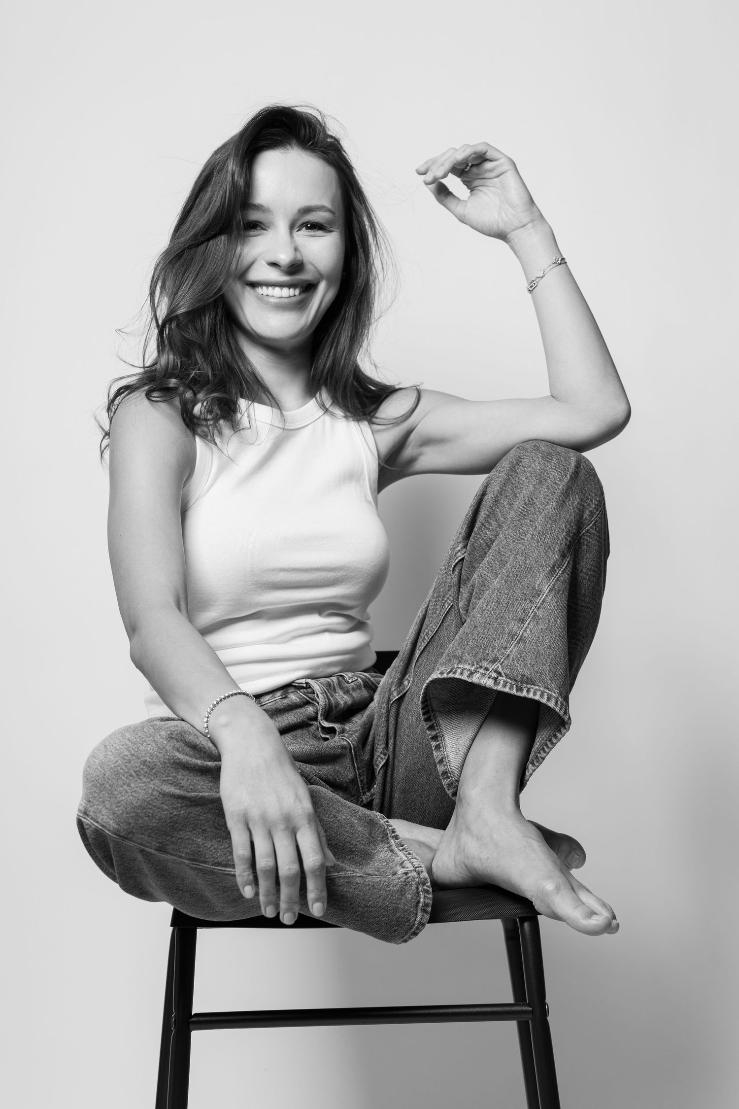 Smiling woman sitting cross-legged on stool in white tank and jeans, black and white studio portrait.