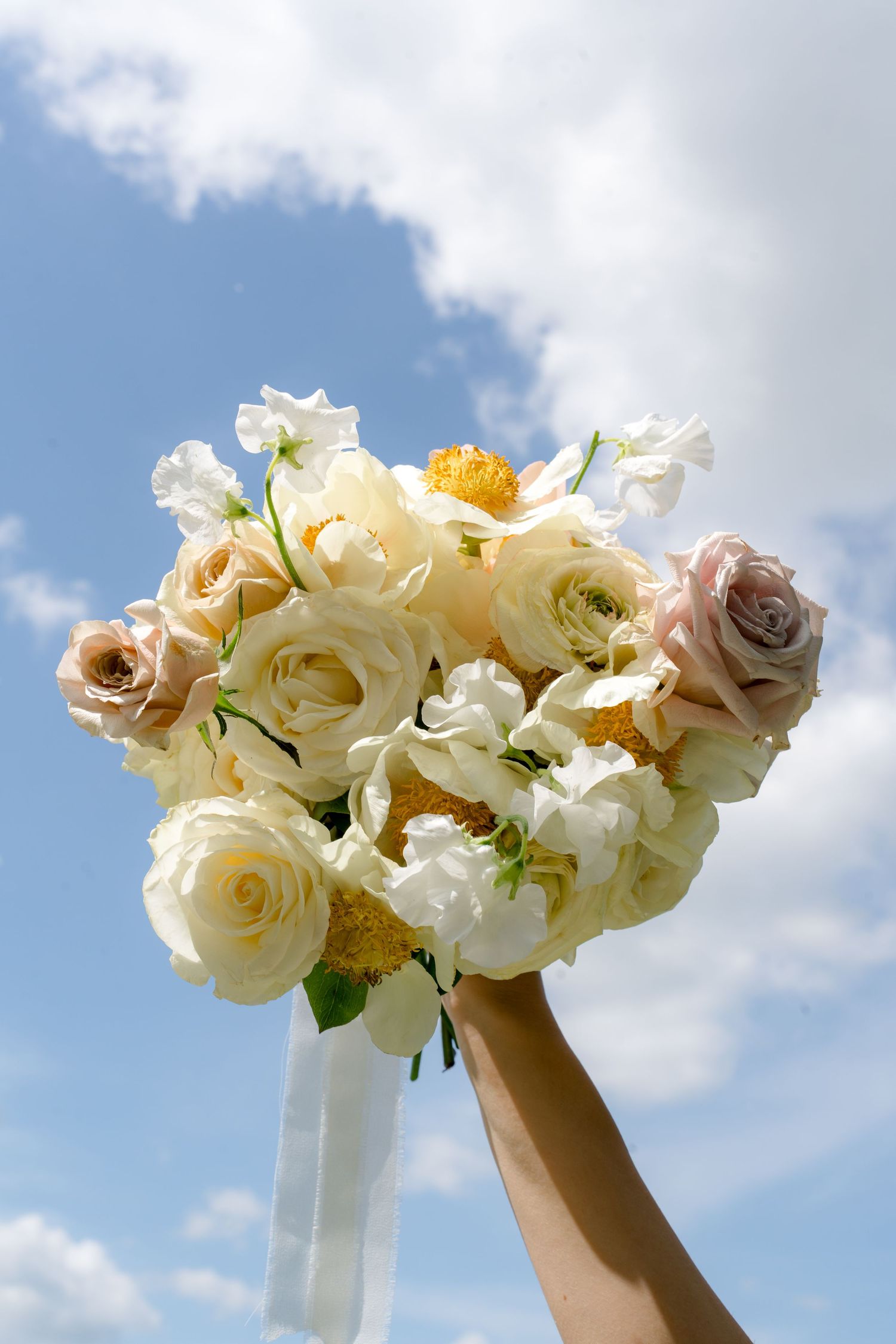 Bridal bouquet of white, cream, and blush roses held up against a blue cloudy sky.