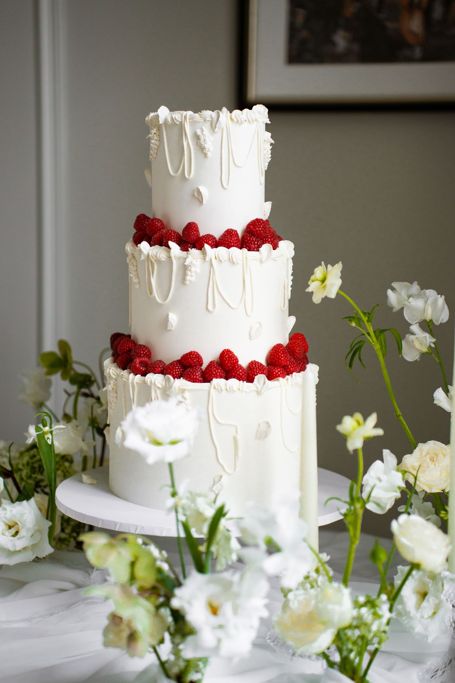 Three-tier white wedding cake with raspberry garnish, dripping white chocolate, and pearl details surrounded by white flowers.