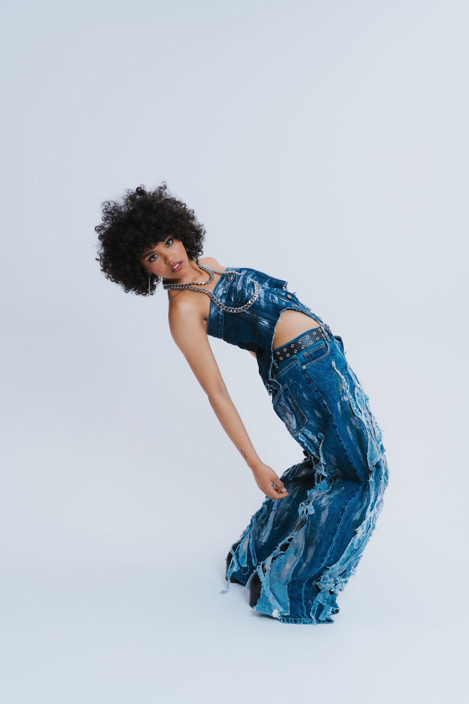 Woman with curly afro hair in denim outfit bending forward gracefully on light blue background.