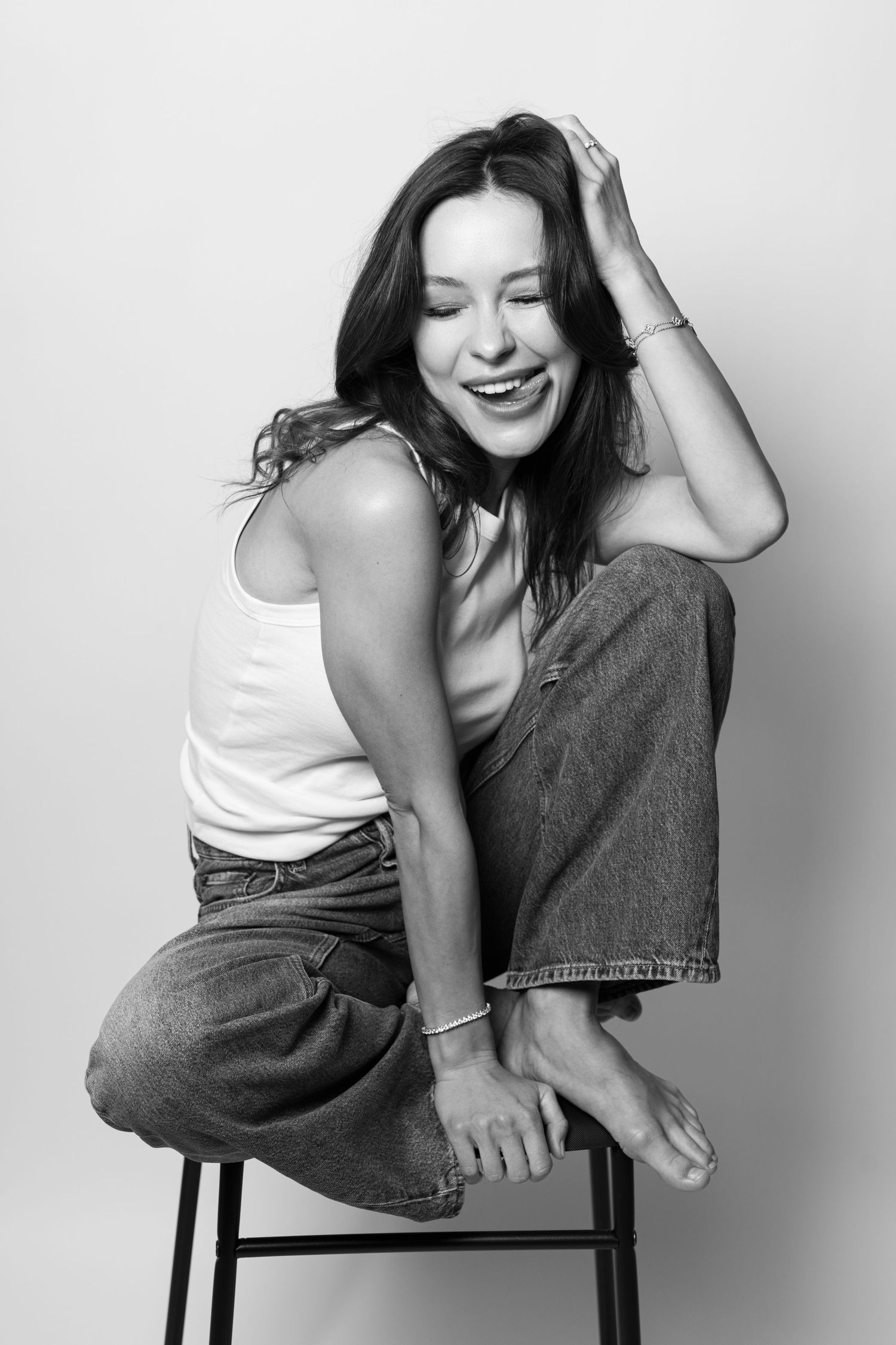 Black and white photo of laughing woman in tank top and jeans sitting on stool.