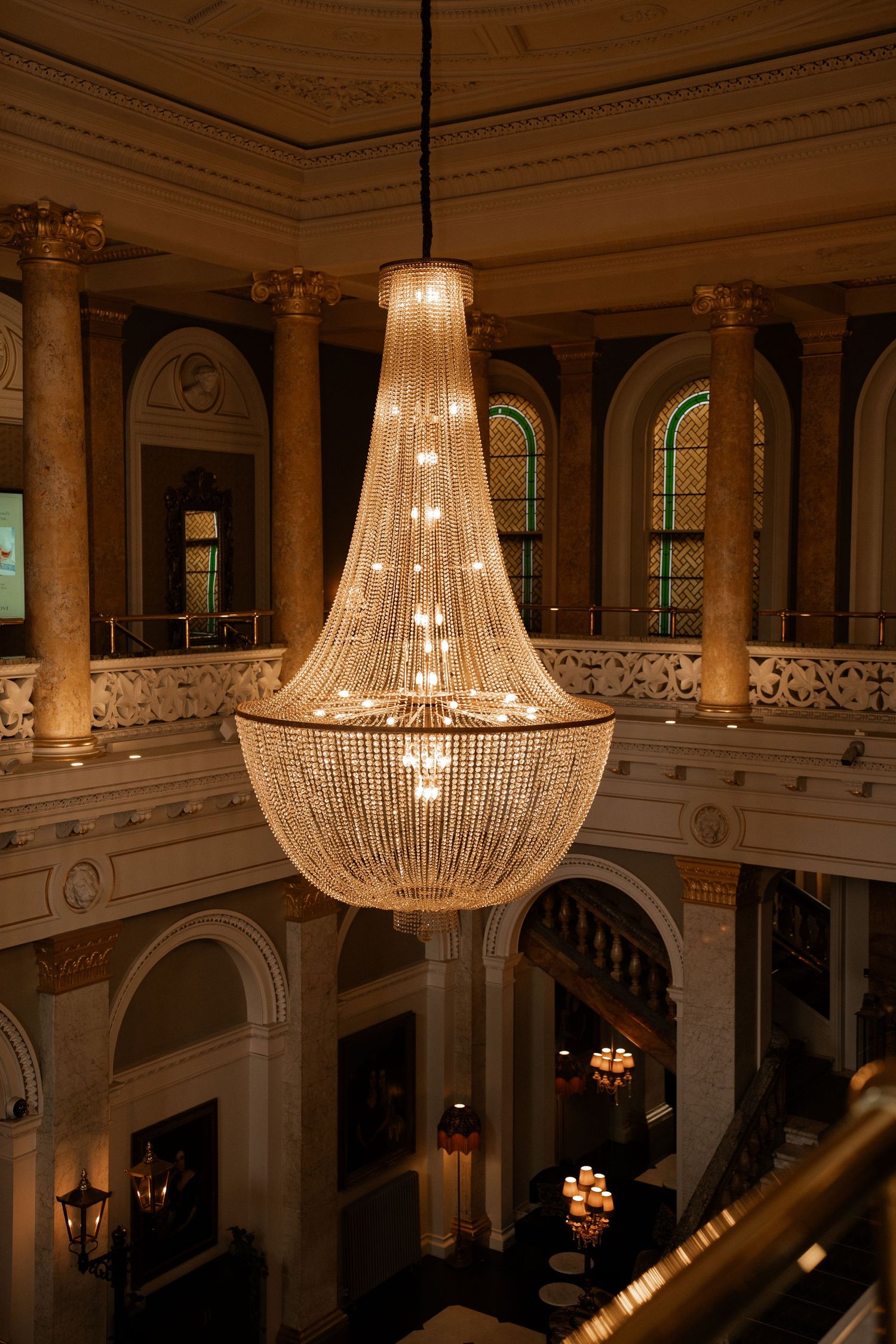 Elegant crystal chandelier hanging in a grand neoclassical hotel lobby with ornate columns and arched windows.
