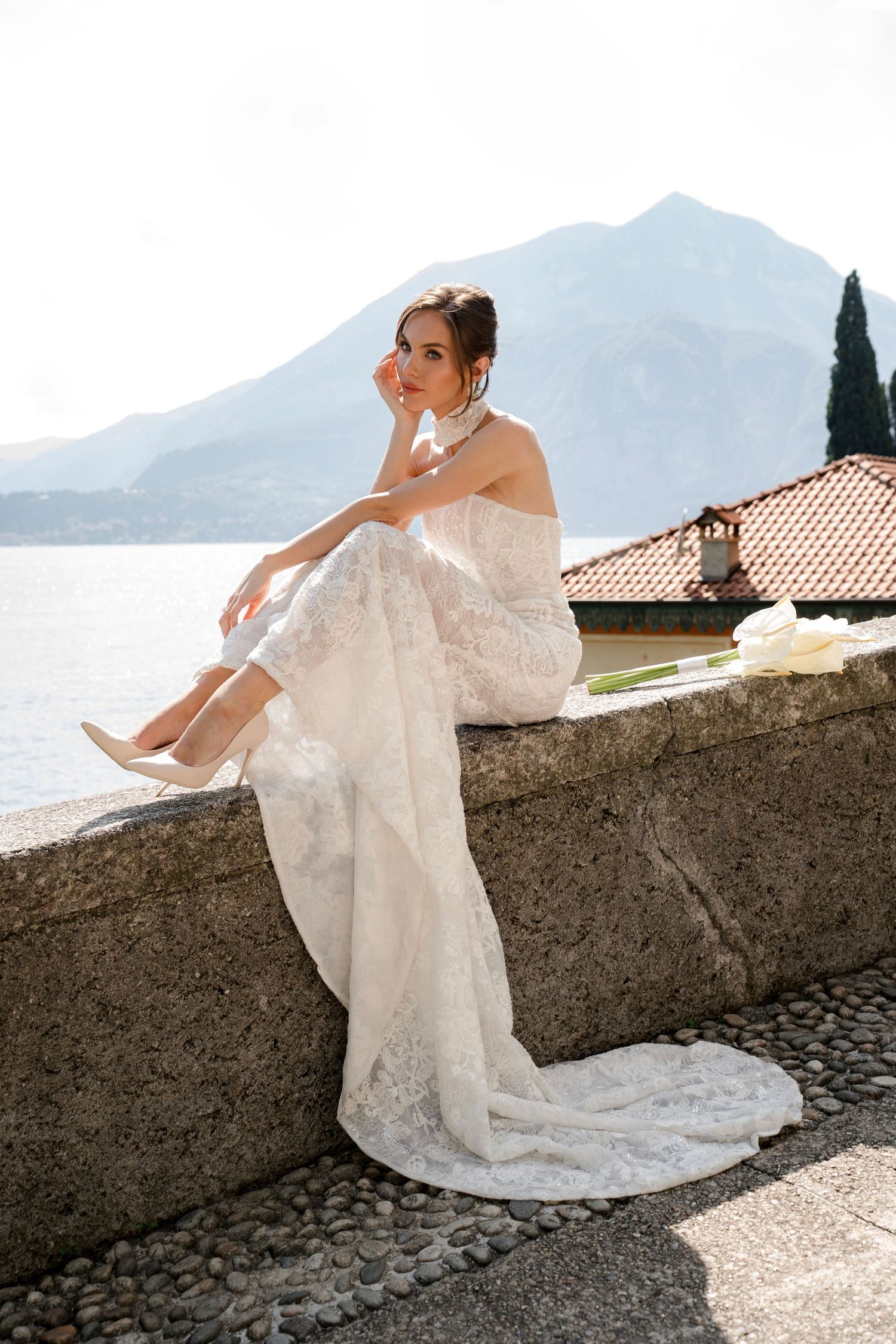 Bride in white lace gown sitting on stone wall with mountain lake backdrop at Italian villa.