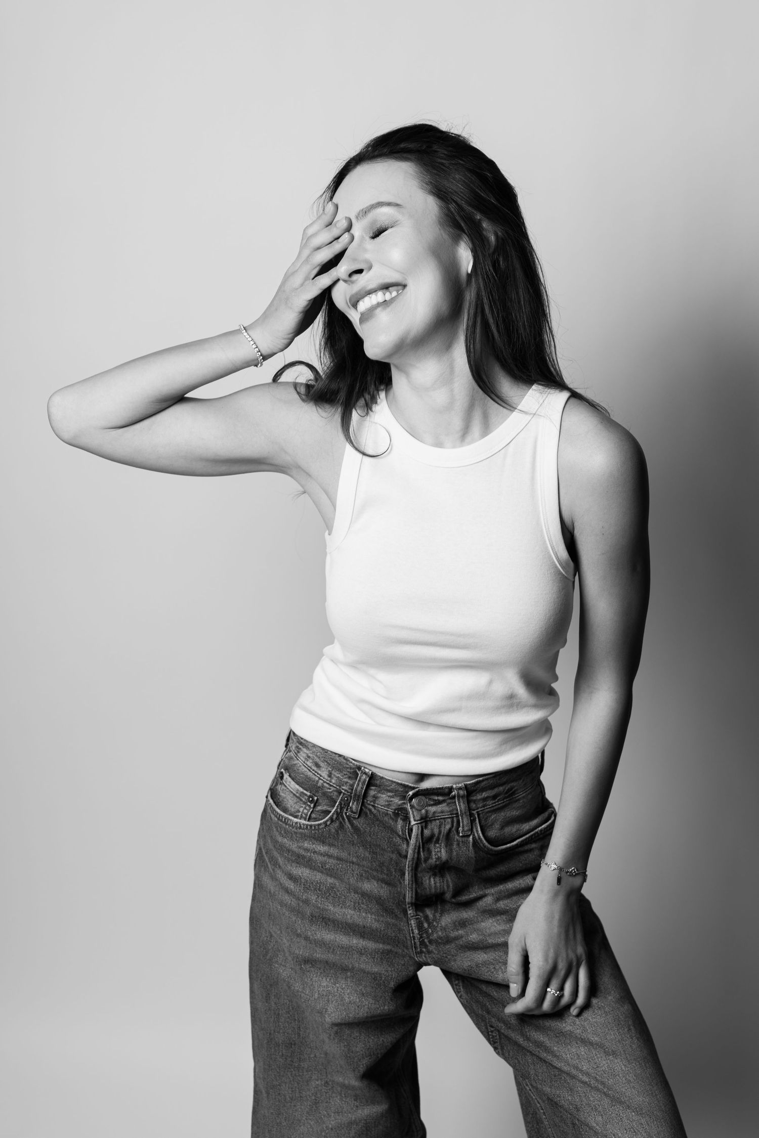 Laughing woman in white tank top and jeans covering face with hand in black and white studio photo.