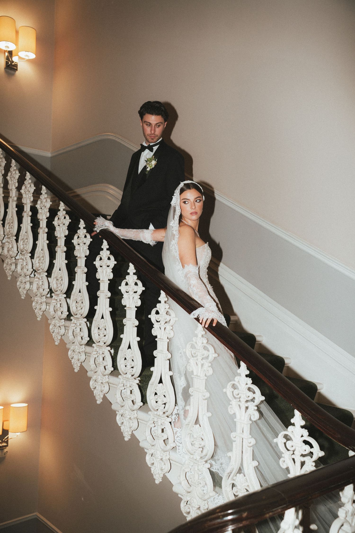 Bride in lace gown with veil and gloves posing with groom on elegant white staircase.