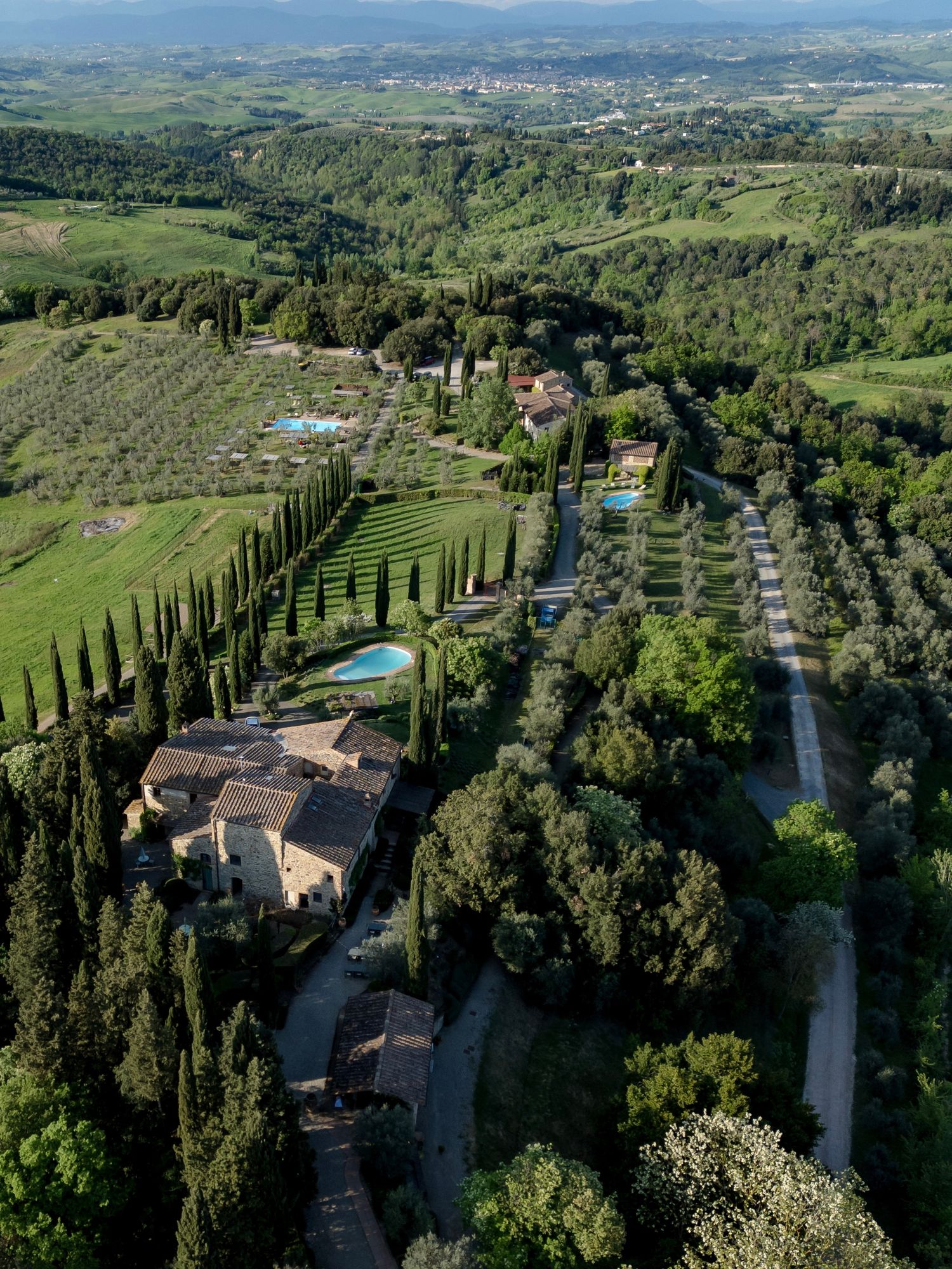 Aerial view of a Tuscan estate surrounded by cypress trees, olive groves, and rolling green countryside with pools.