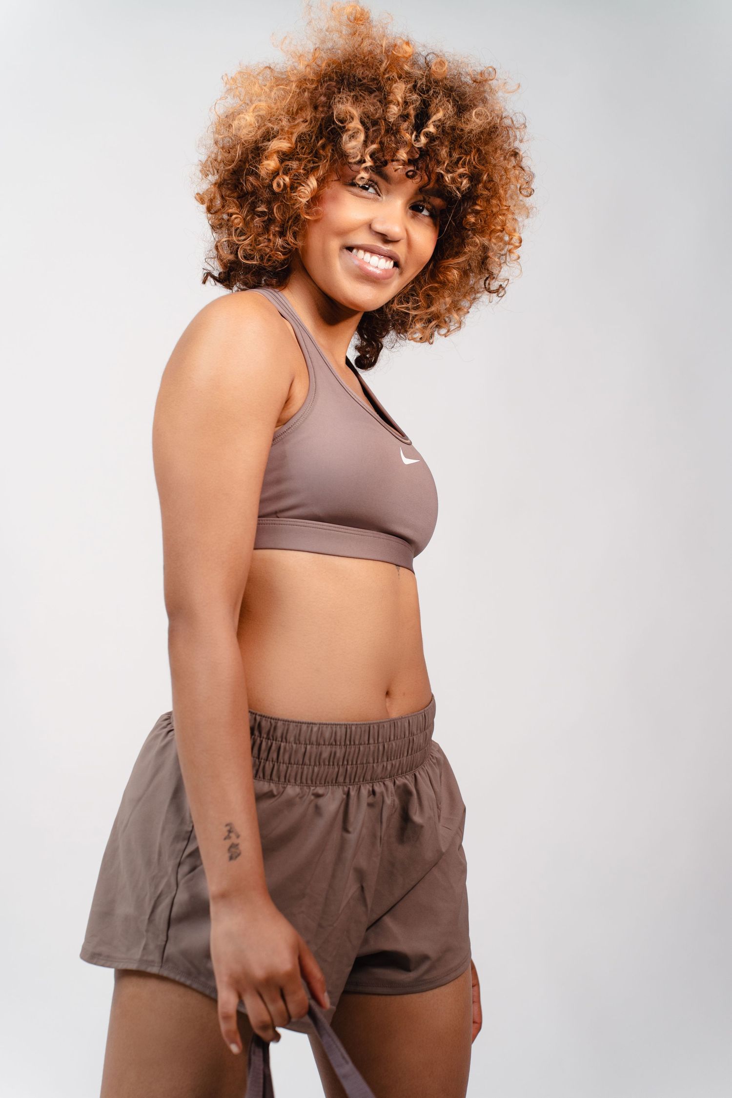 Smiling curly-haired woman in taupe Nike sports bra and shorts posing on white background.