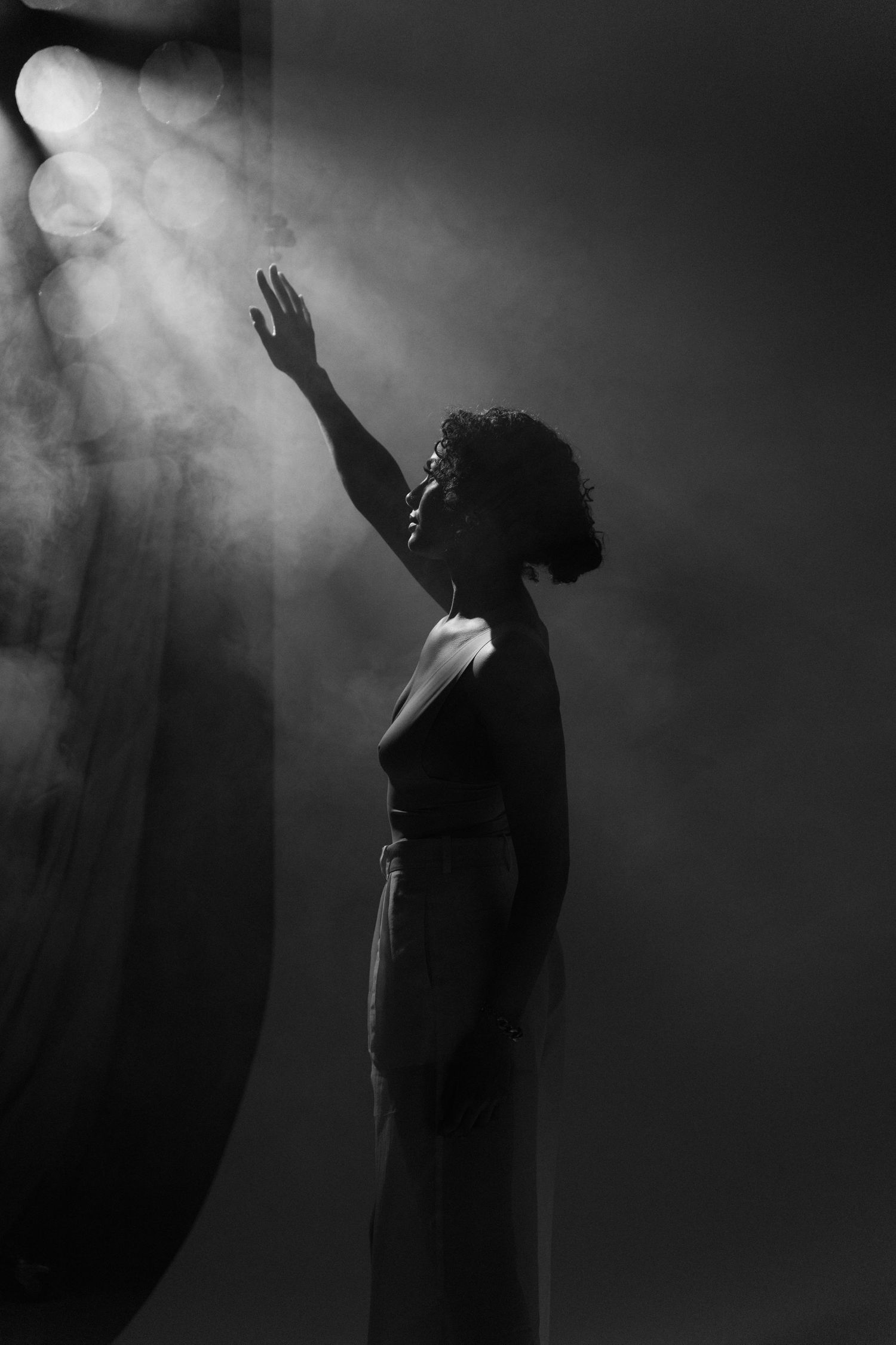 Dramatic black and white silhouette of woman reaching up toward light rays in smoky studio setting.