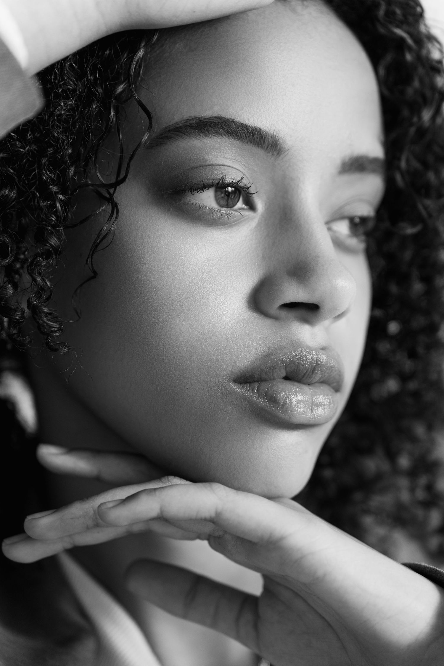 Black and white close-up portrait of a curly-haired woman with hand near chin in soft dramatic lighting.