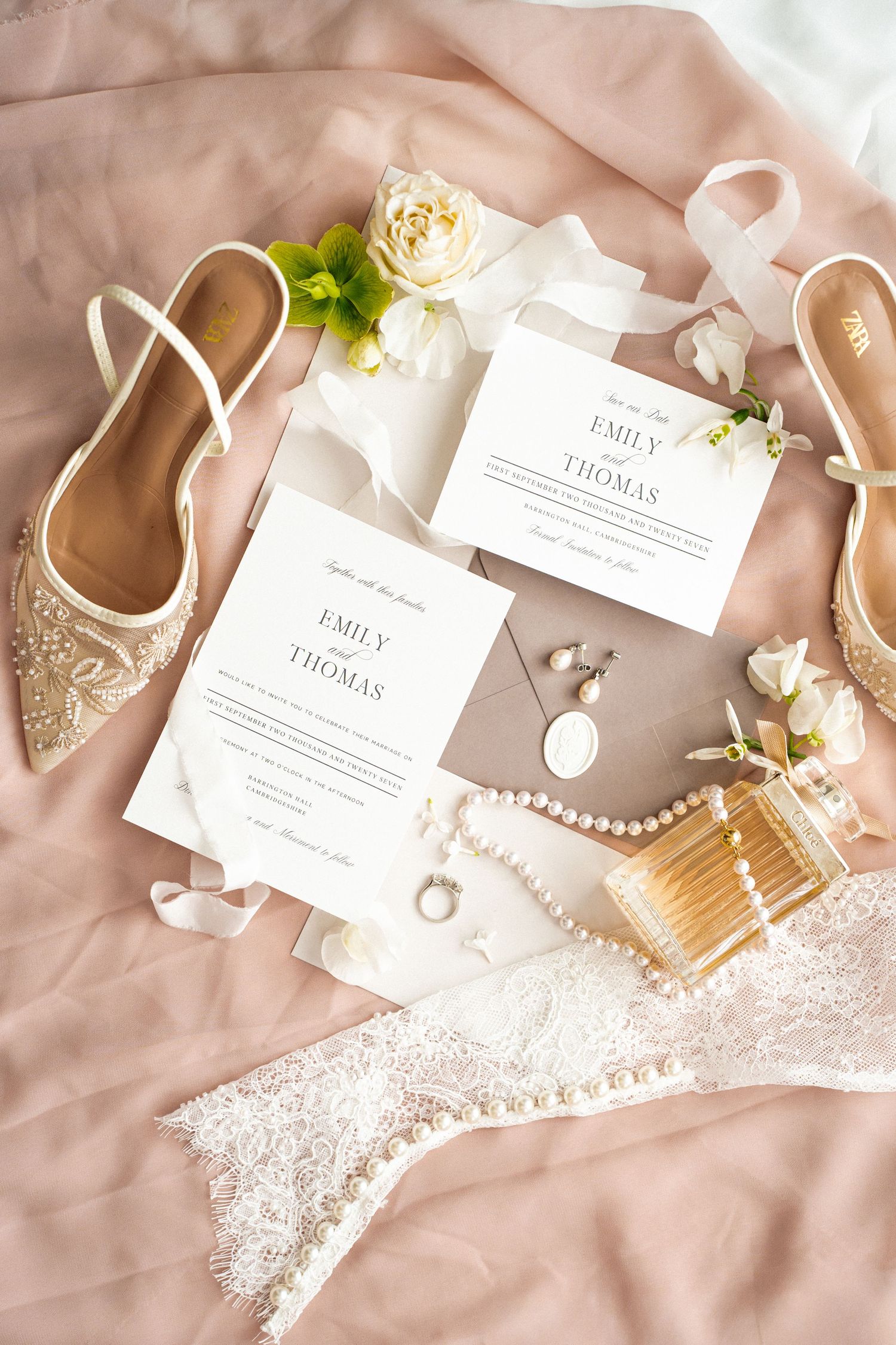 Elegant wedding flat lay with ivory invitations, pearl necklace, perfume, lace details, and embellished bridal shoes on blush fabric.