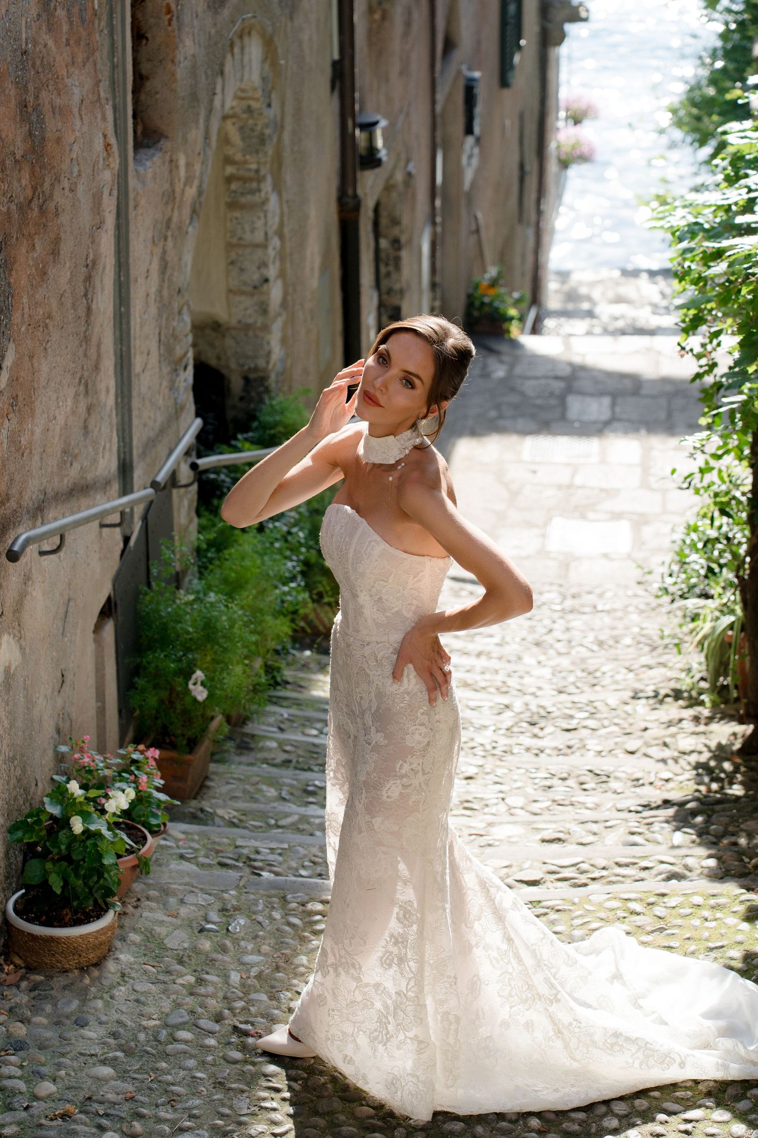Bride in fitted lace wedding gown poses on charming cobblestone alley with stone walls and greenery.