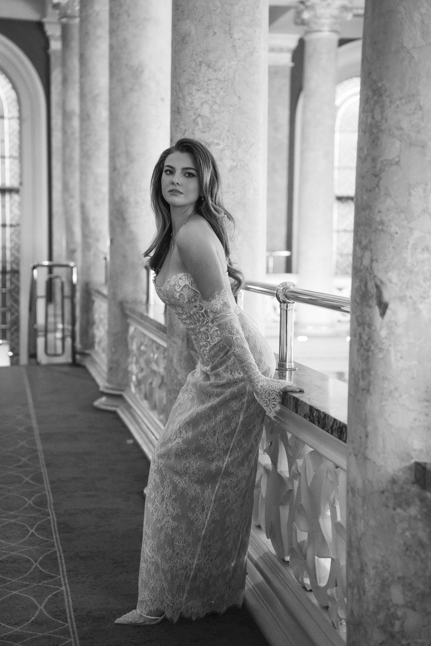 Black and white photo of a woman in a lace gown posing gracefully by ornate marble columns in an elegant hall.
