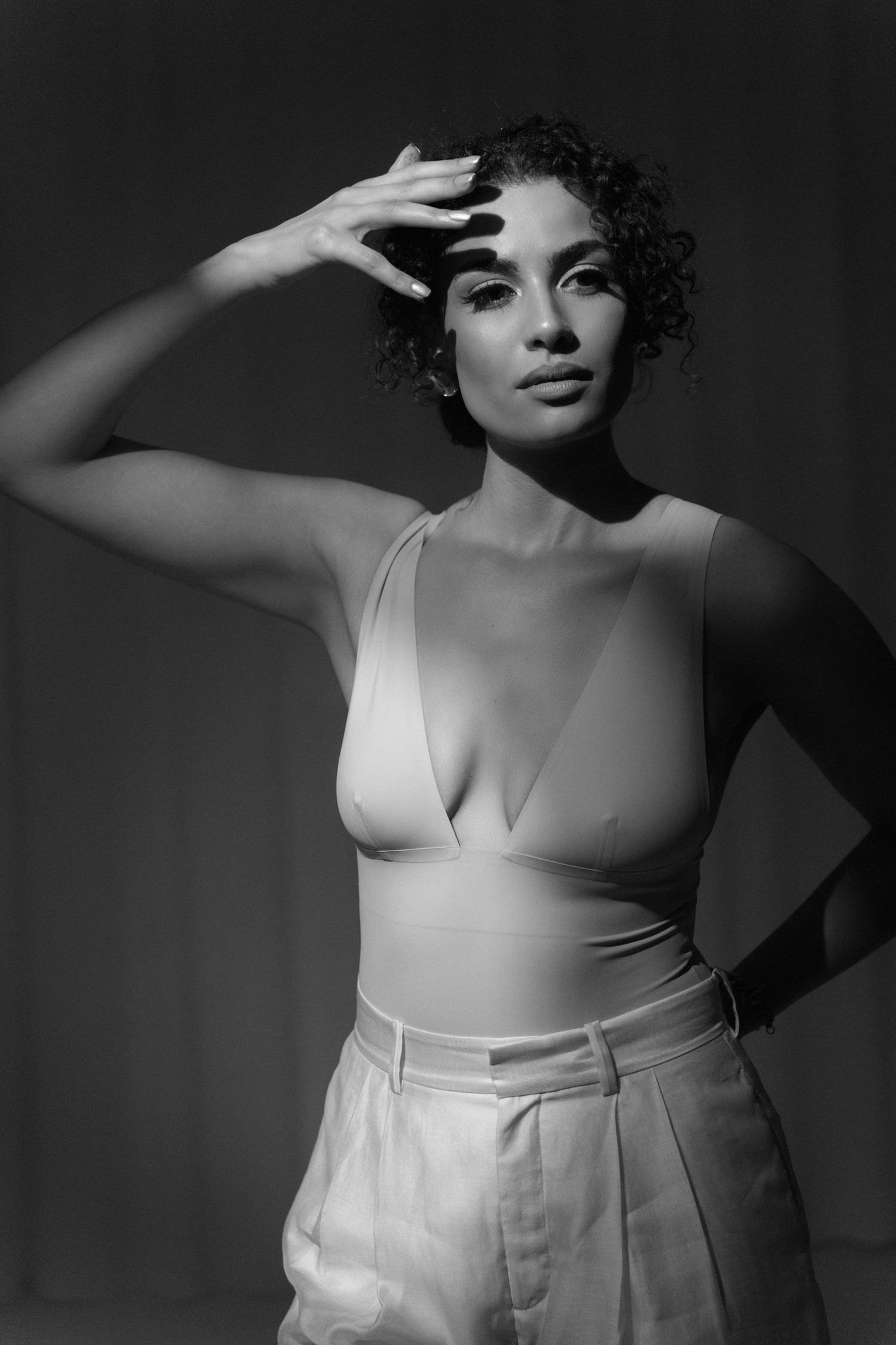 Black and white photo of woman in low-cut bodysuit and wide-leg pants, hand shielding eyes, studio shot.