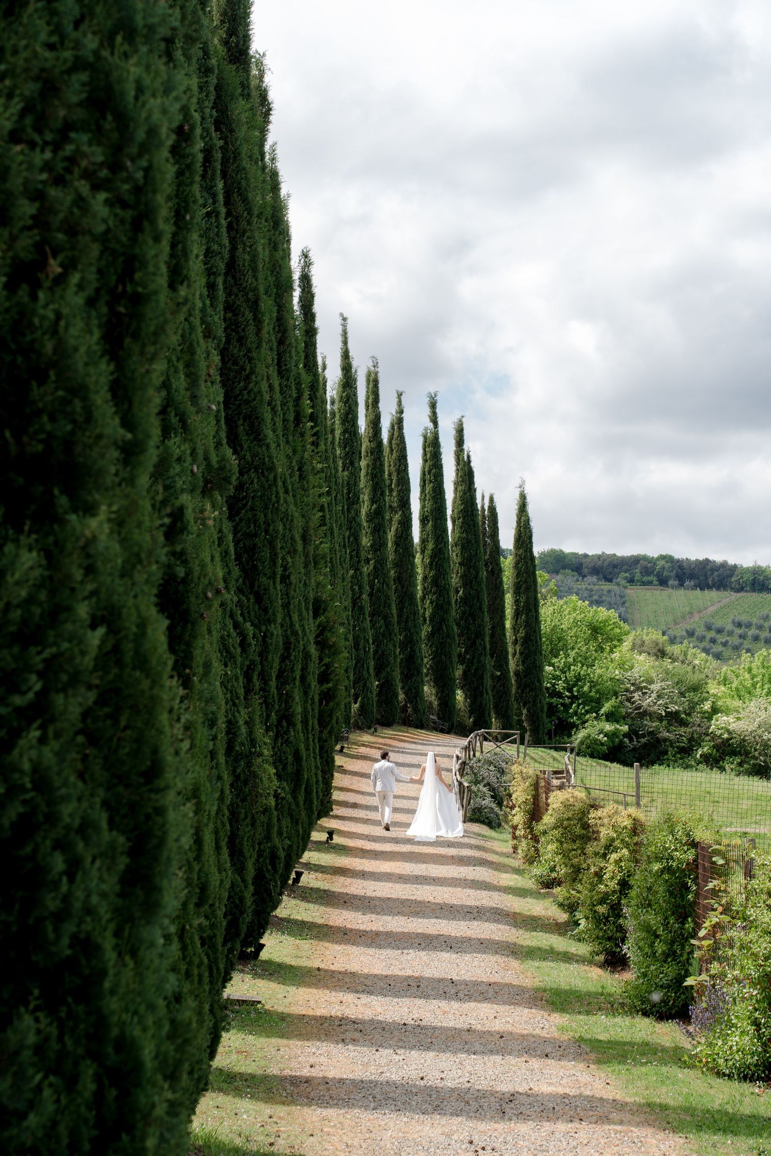 Bride and groom walk along a gravel path lined with tall cypress trees at a Tuscan estate.