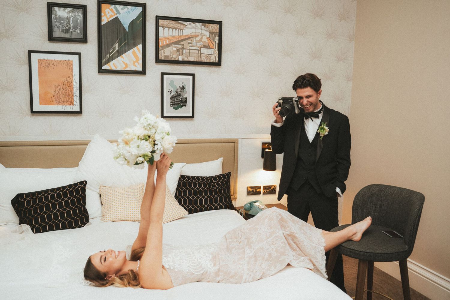 Playful bride in wedding dress lying on hotel bed holding bouquet while groom photographs her with vintage camera.