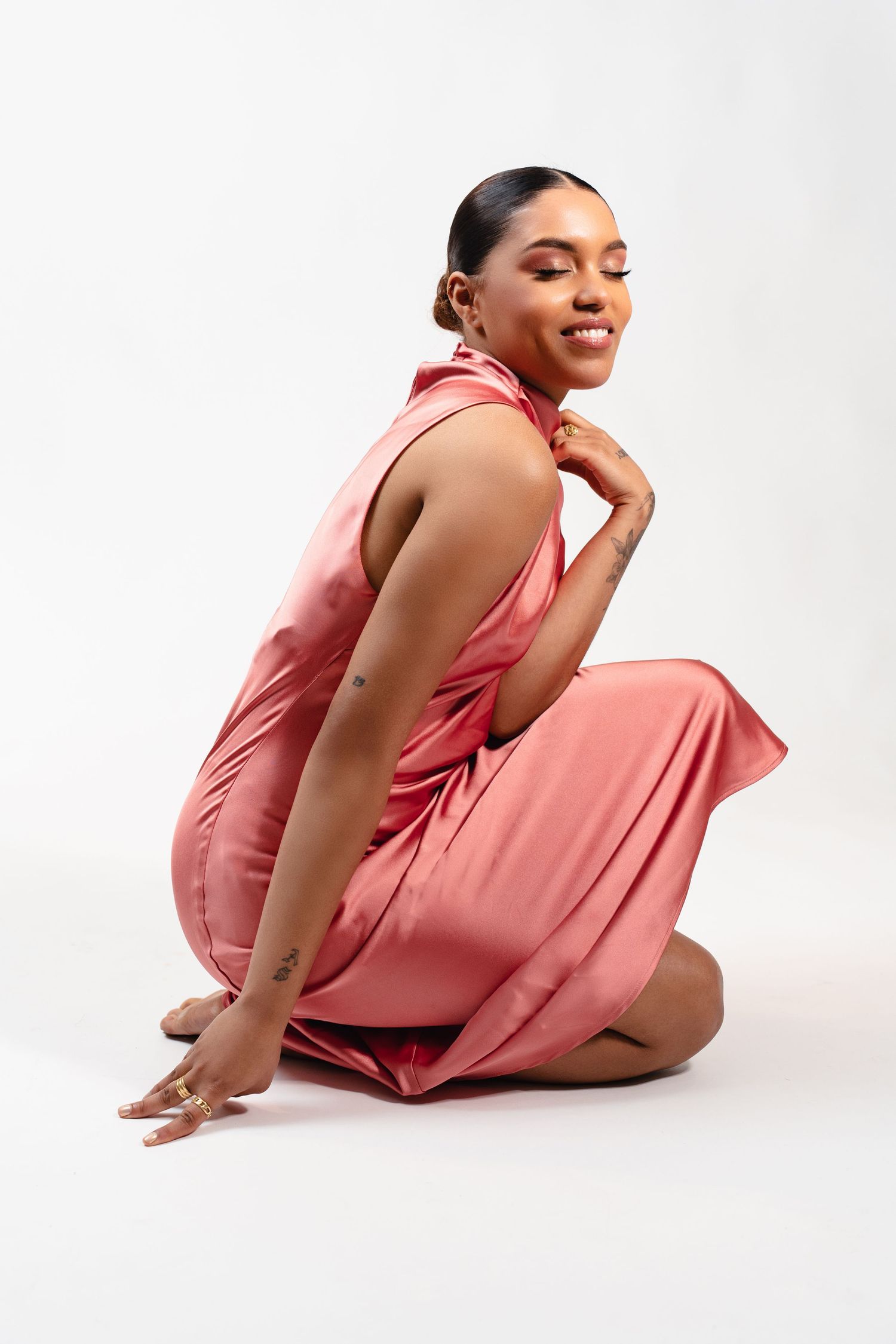 Woman in a flowing pink satin halter dress sitting on floor, smiling over her shoulder against white background.