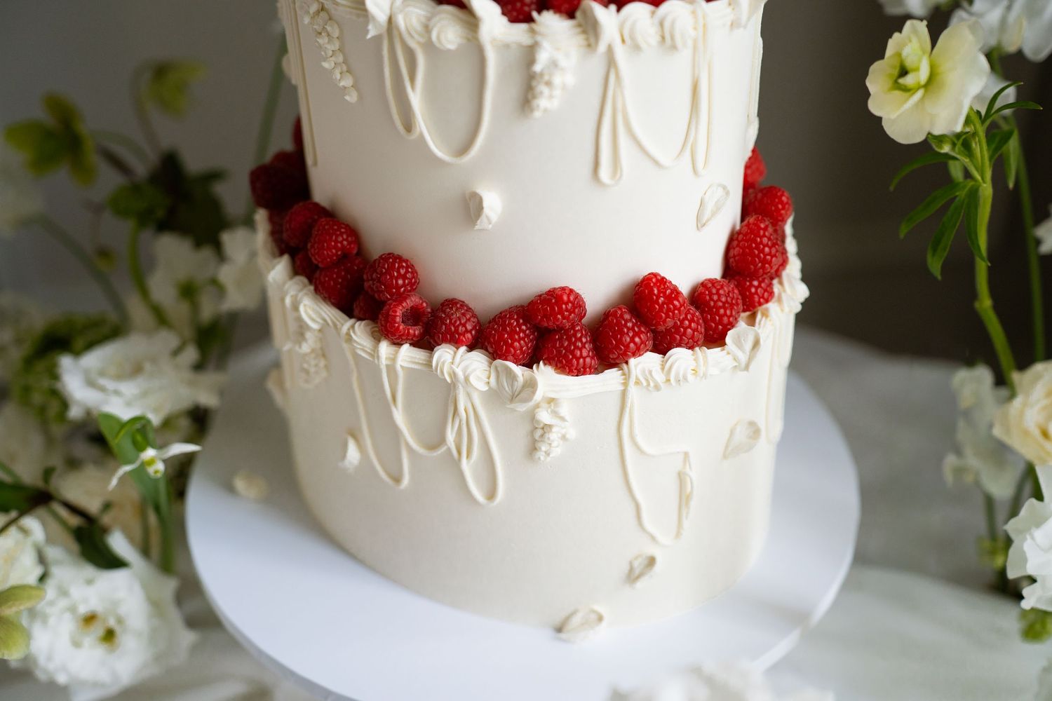 Elegant two-tier white wedding cake with raspberry border and white chocolate drizzle, surrounded by fresh flowers.
