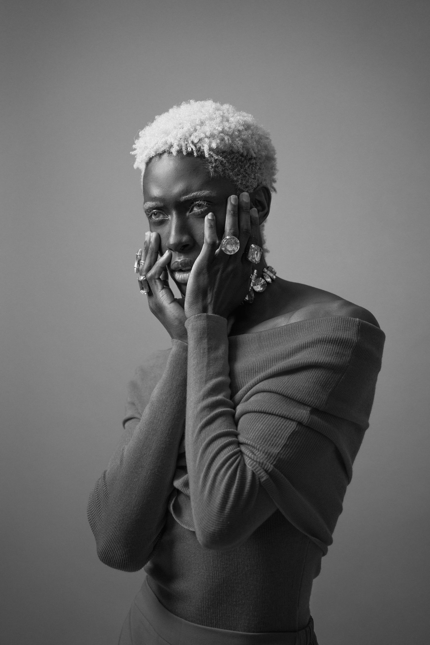 Black and white portrait of a person with short bleached hair posing with hands near face and statement rings.