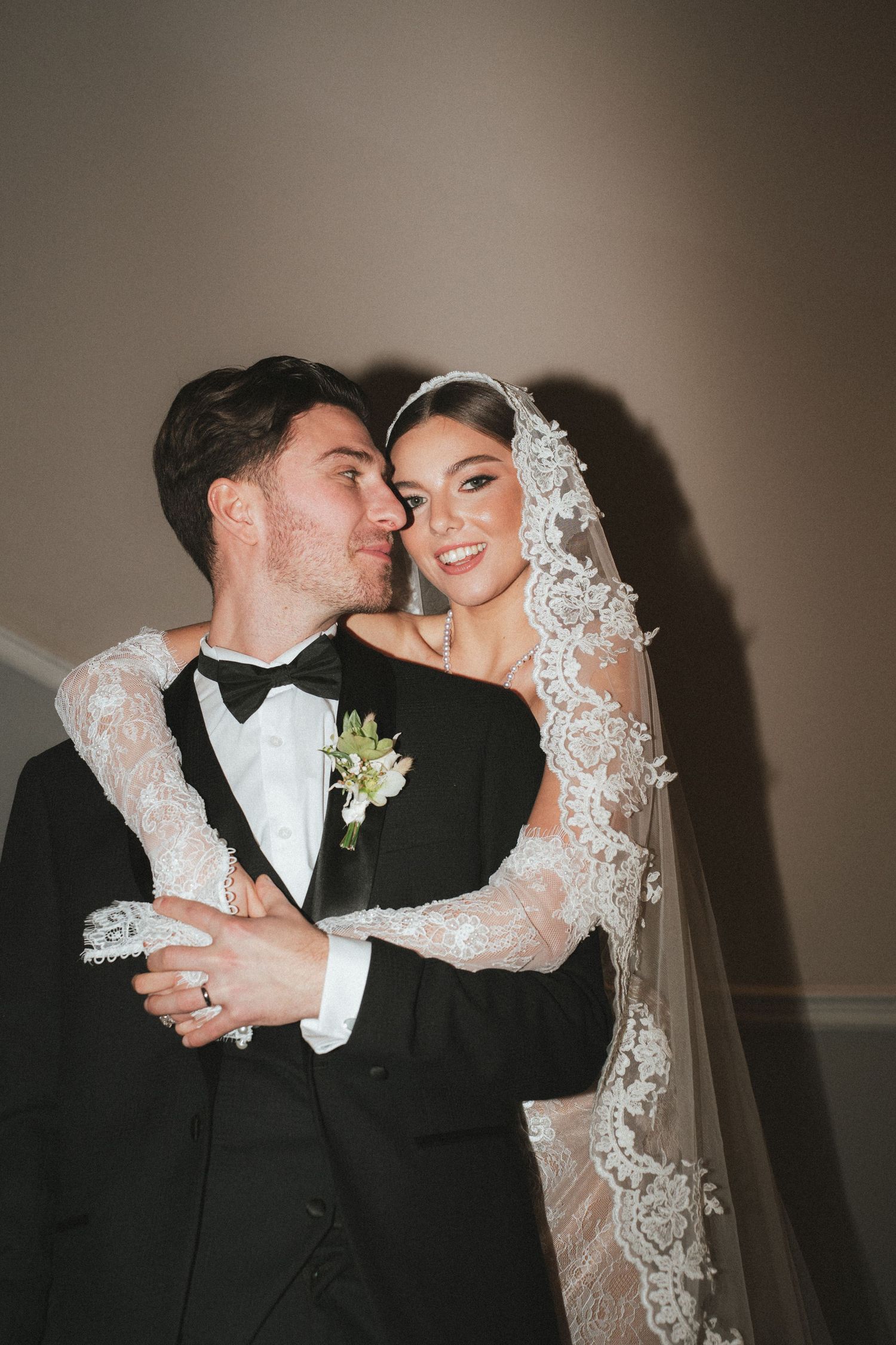 Bride in lace wedding gown and veil embracing smiling groom in black tuxedo indoors.