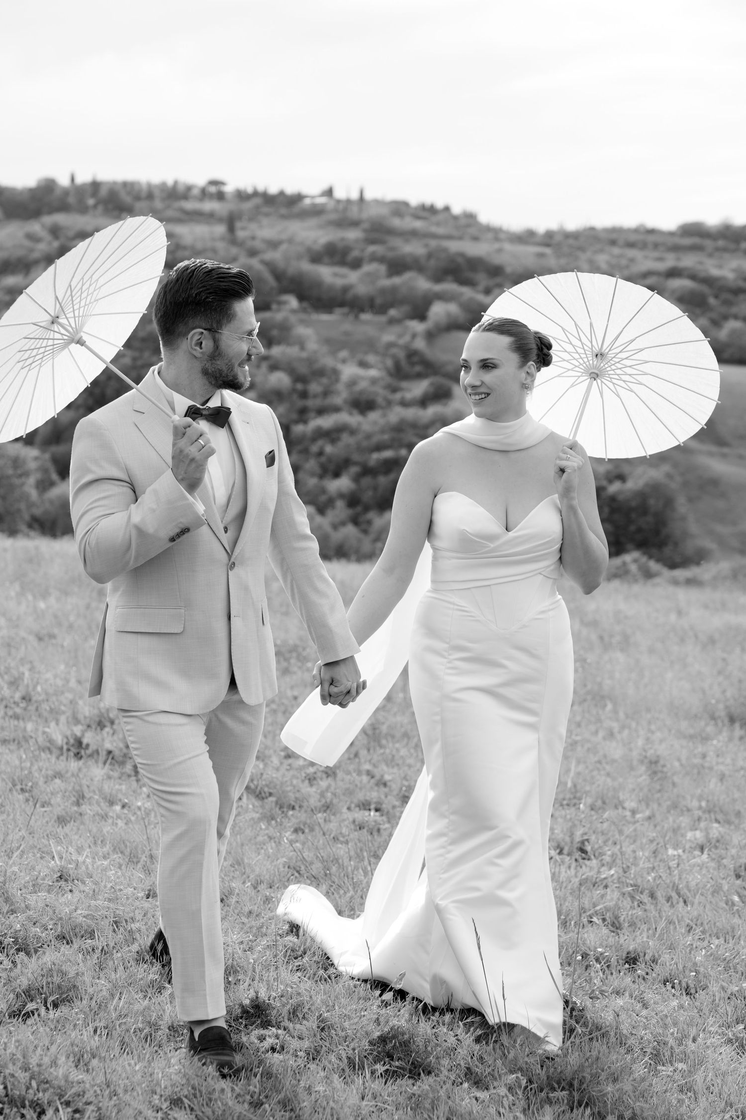 Black and white photo of smiling bride and groom holding parasols in a scenic countryside field.