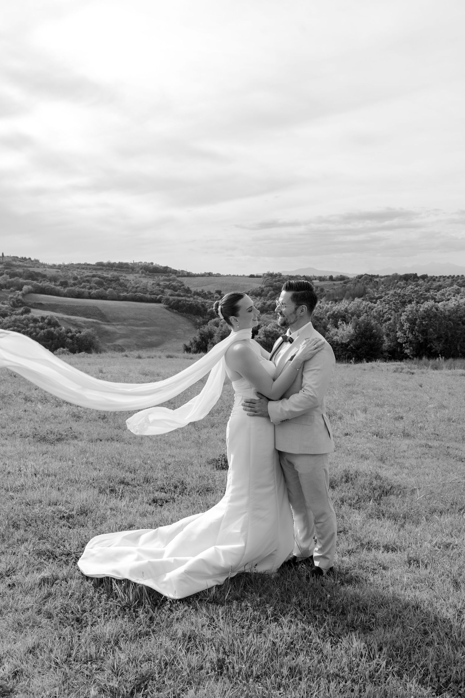 Black and white photo of bride and groom embracing in a field, veil flowing in the wind with rolling hills behind.