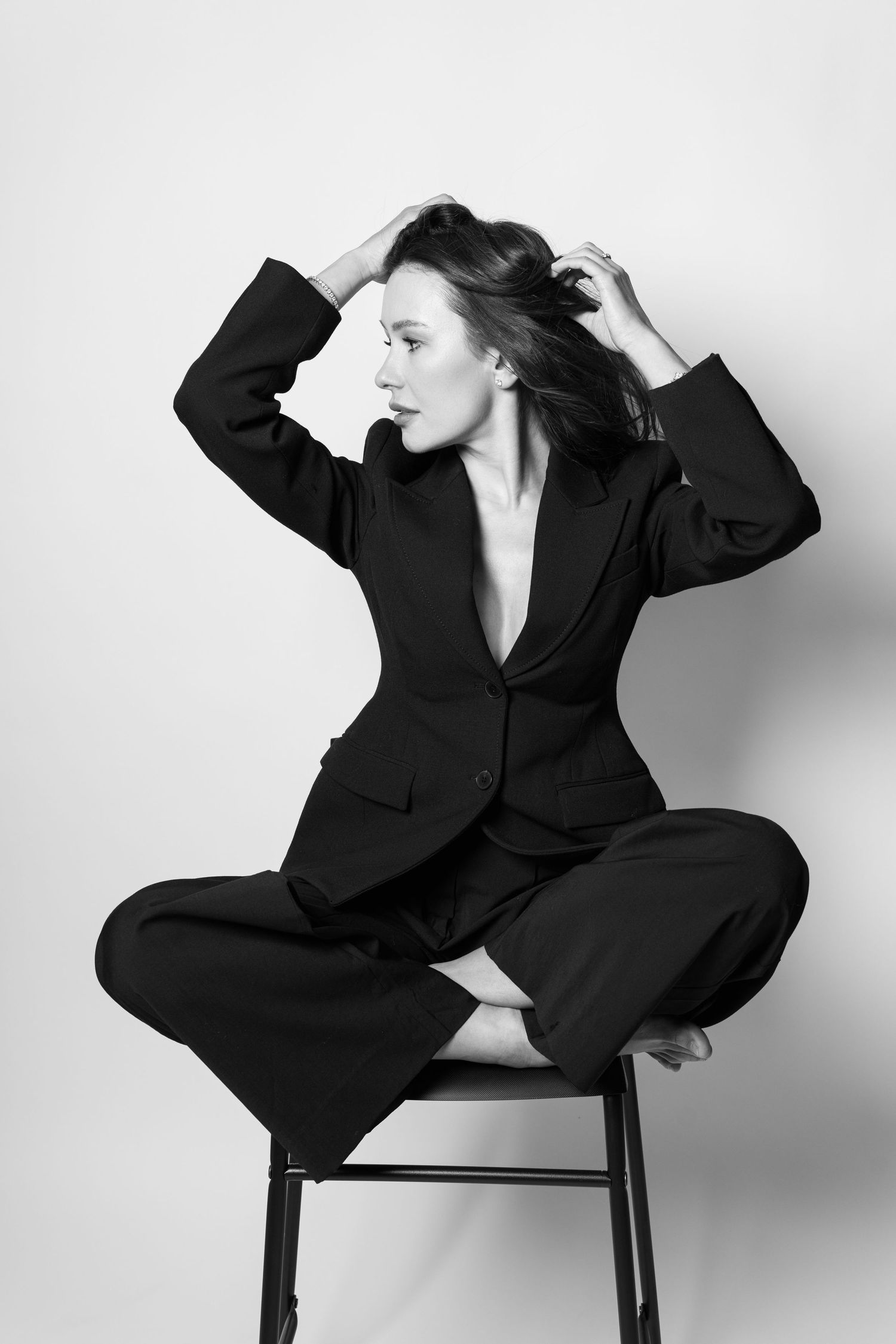 Woman in black suit sitting on stool styling hair in black and white studio portrait.