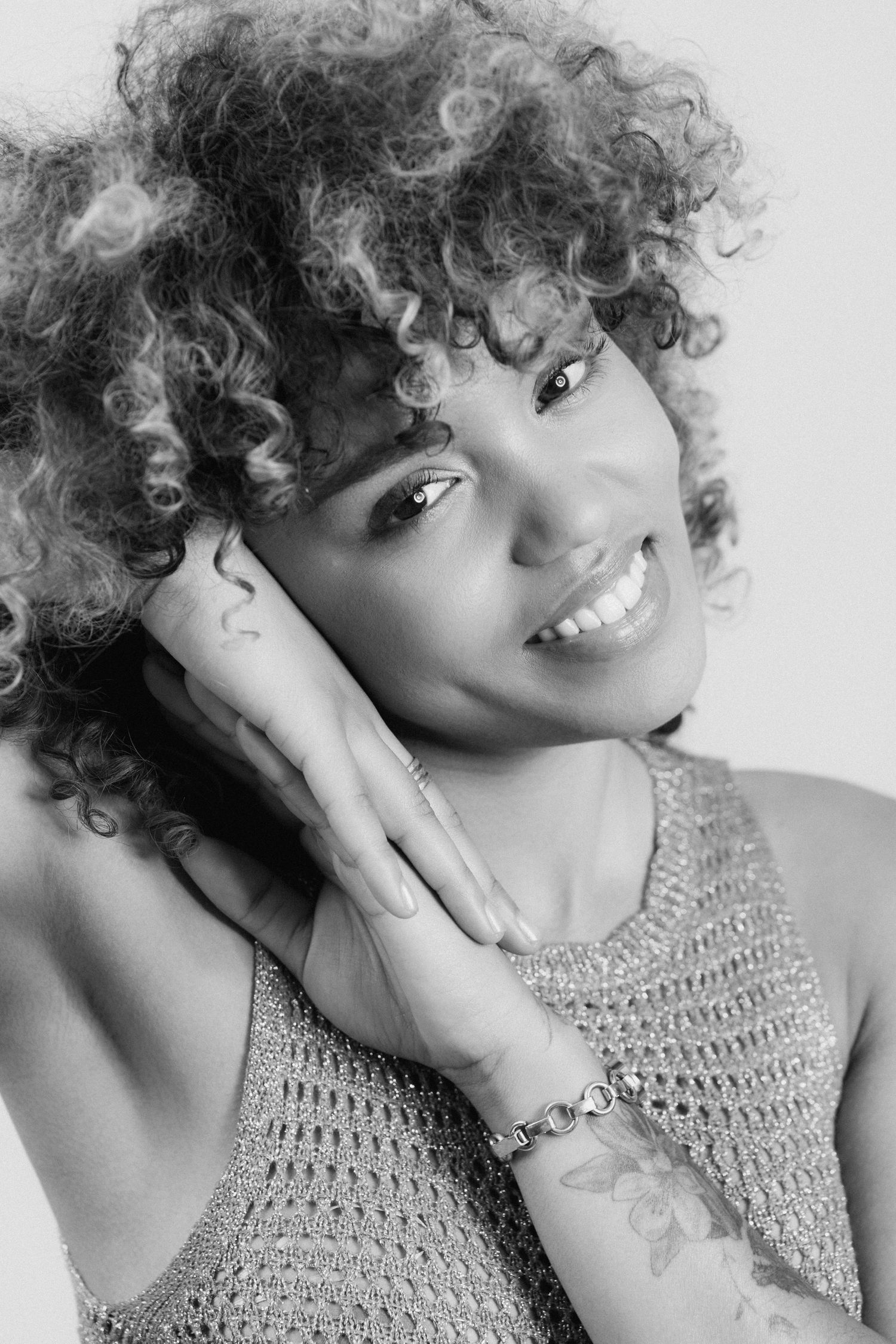 Black and white portrait of a smiling woman with curly hair, hands near face, wearing a bracelet and showing tattoos.