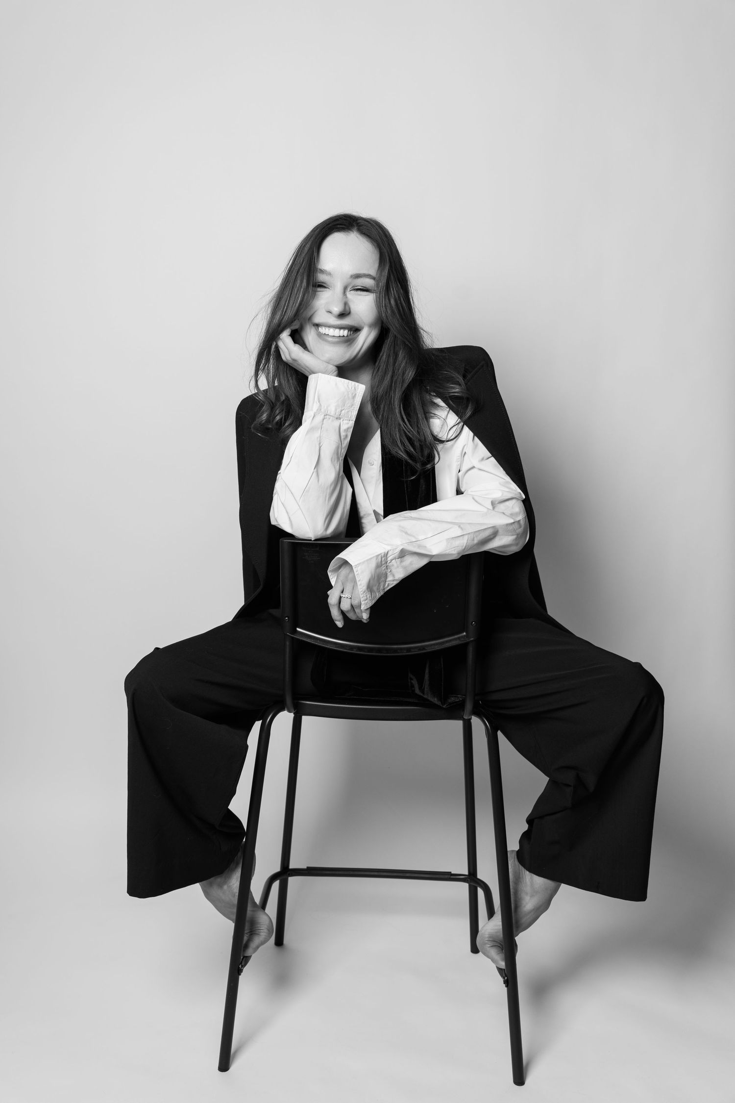 Woman in black suit and white shirt sitting on stool laughing in black and white studio photo.