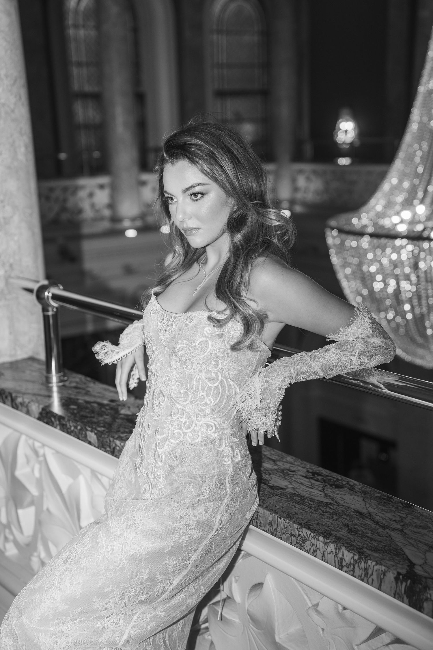 Black and white portrait of an elegant bride in lace gown and gloves leaning on a marble railing.