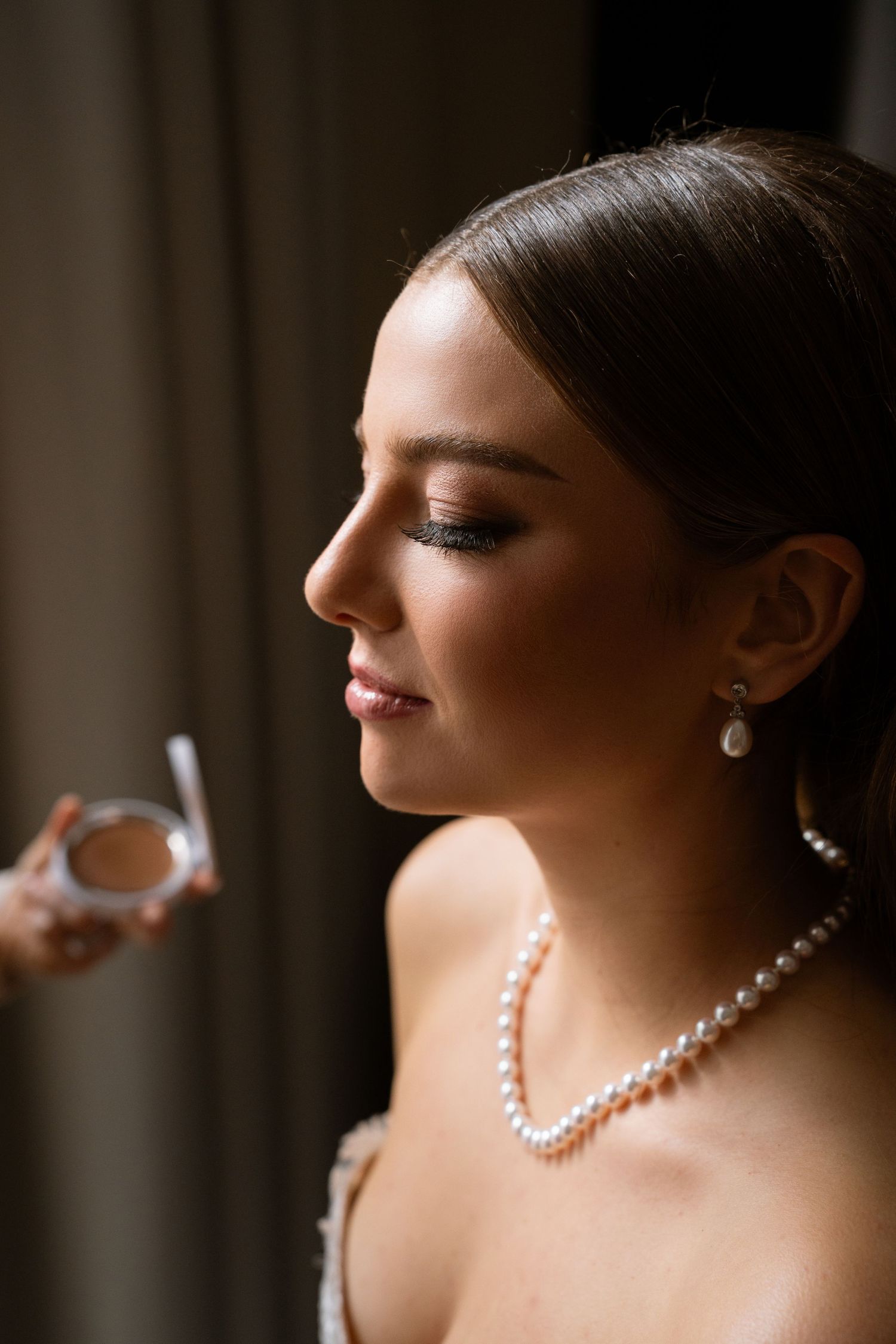 Beautiful bride in profile view wearing pearl necklace and earrings during wedding makeup application.