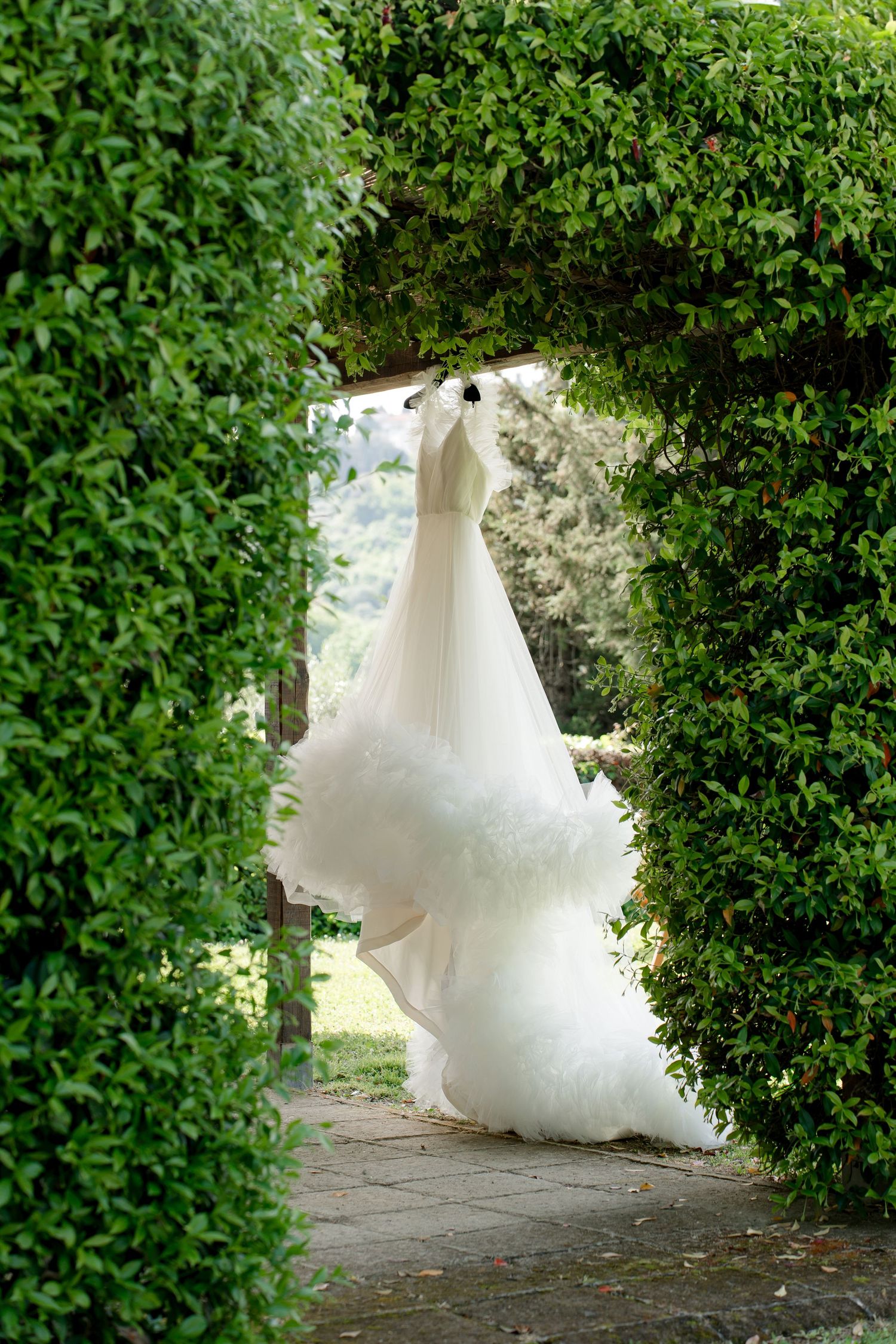 Elegant white wedding gown with tulle skirt hanging in a lush green garden archway.