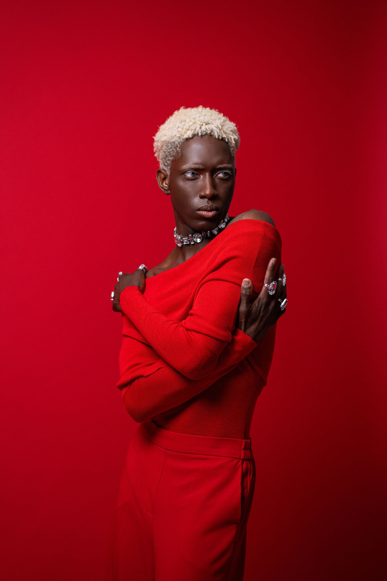 Striking Black woman in red off-shoulder dress with blonde hair and silver jewelry on red background.