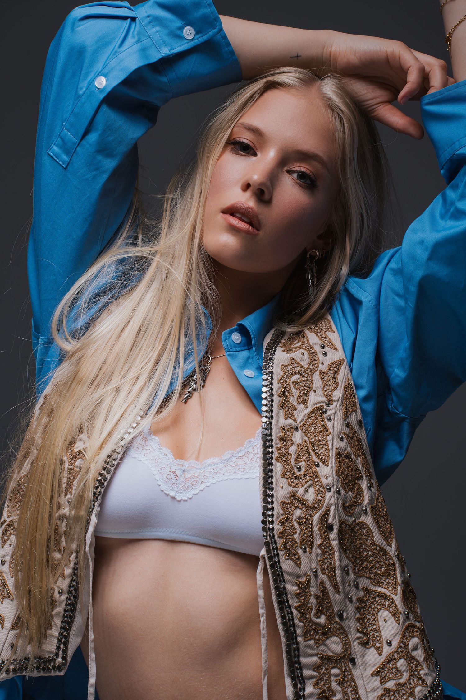 Blonde model posing in blue shirt and embroidered vest with white bralette in studio.