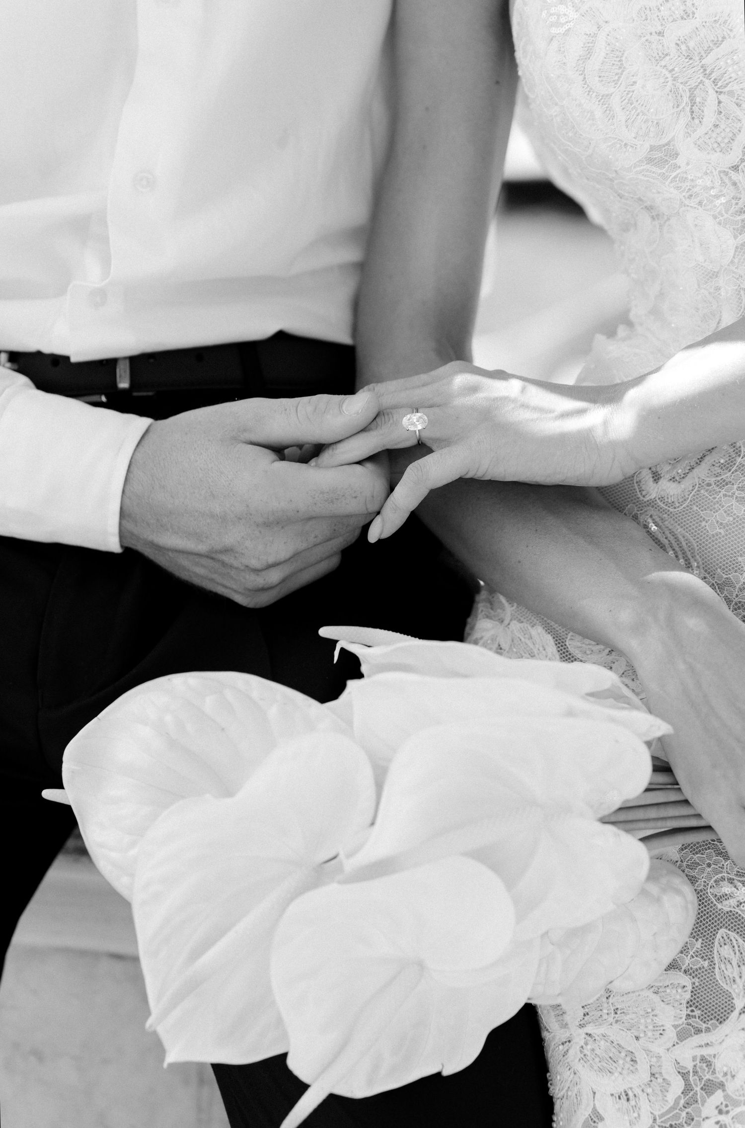 Black and white photo of bride and groom exchanging rings, with white calla lily bouquet in foreground.