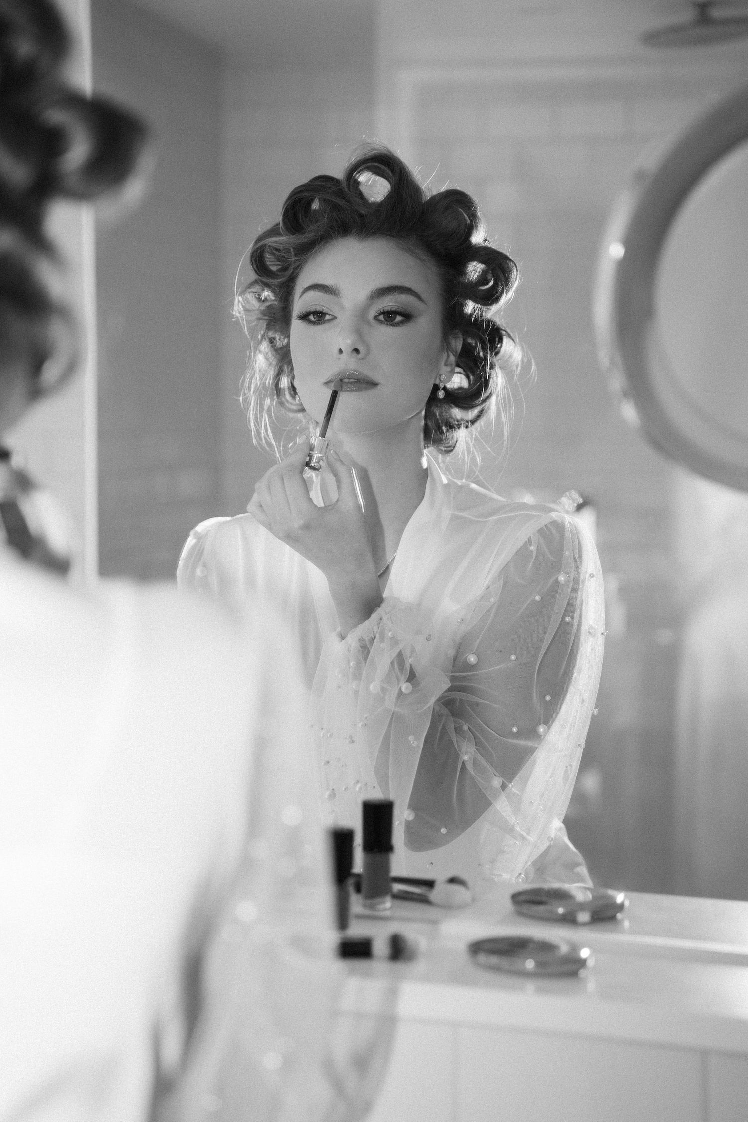 Black and white photo of bride with hair curlers applying lipstick at vanity mirror in sheer white robe.
