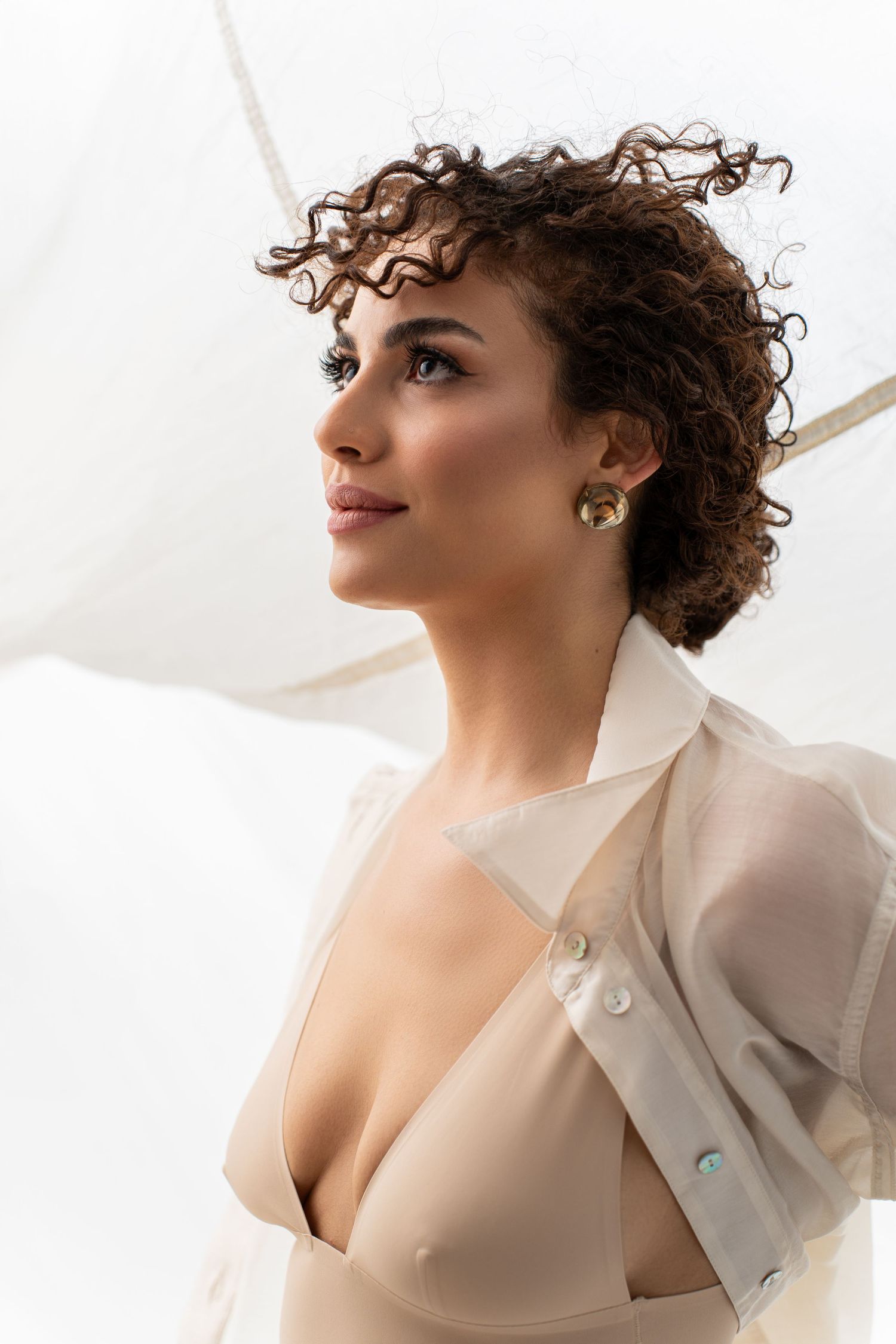 Woman with curly hair wearing sheer white blouse and beige bra, holding umbrella, looking away.