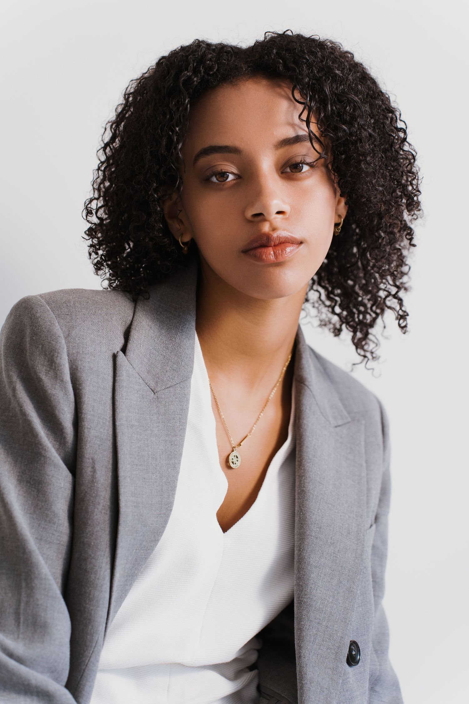 Young woman with curly hair wearing a gray blazer and gold necklace posing against a white background.
