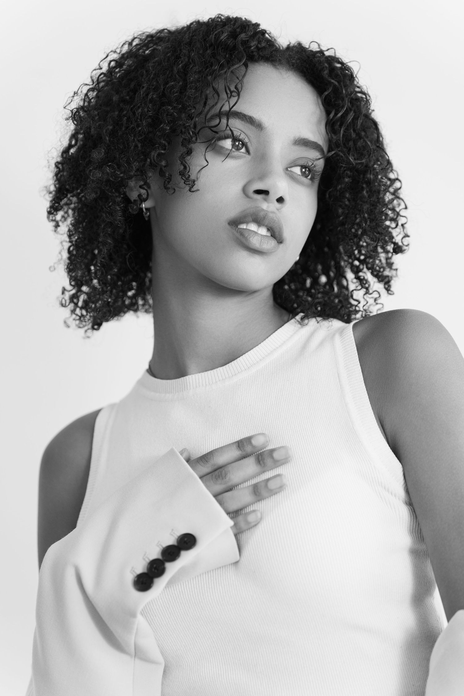Black and white portrait of a curly-haired woman in a white sleeveless top with hand placed on chest.