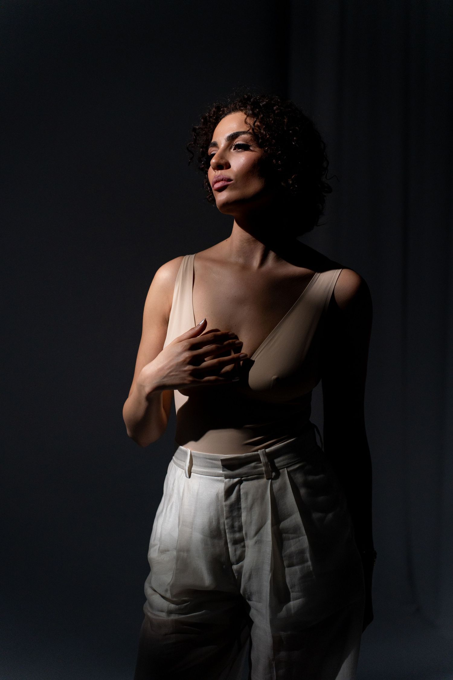 Woman in beige bodysuit and white trousers posing in dramatic shadow lighting against dark background.