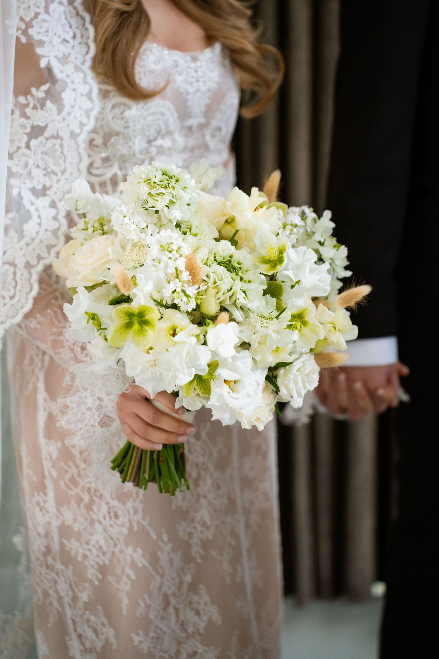 Bride in lace wedding dress holding lush white and green bridal bouquet while holding grooms hand.